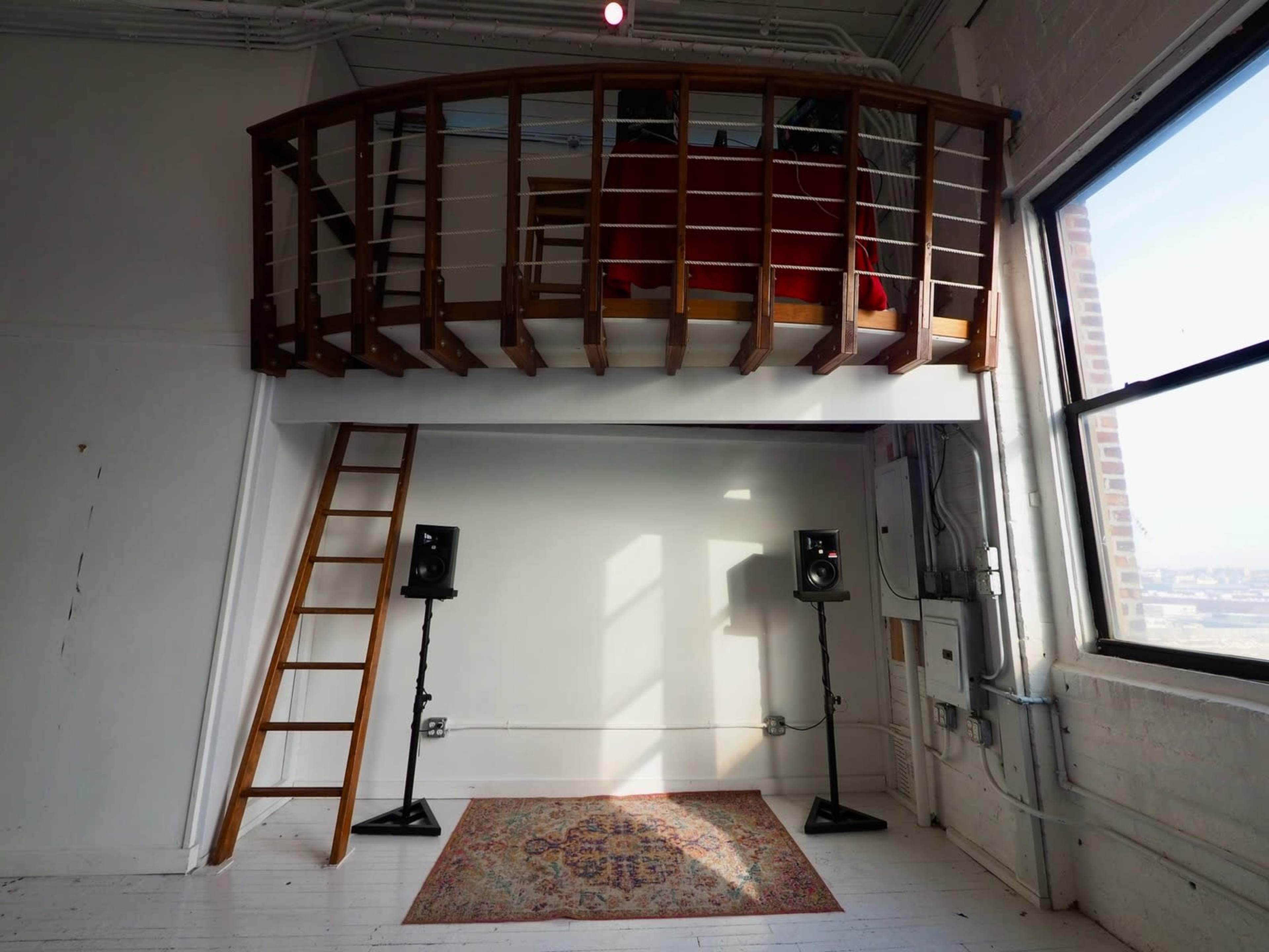 The image shows a minimalist interior space with a wooden loft, a ladder leading to the loft, two speakers on either side, and a patterned rug on the floor.
