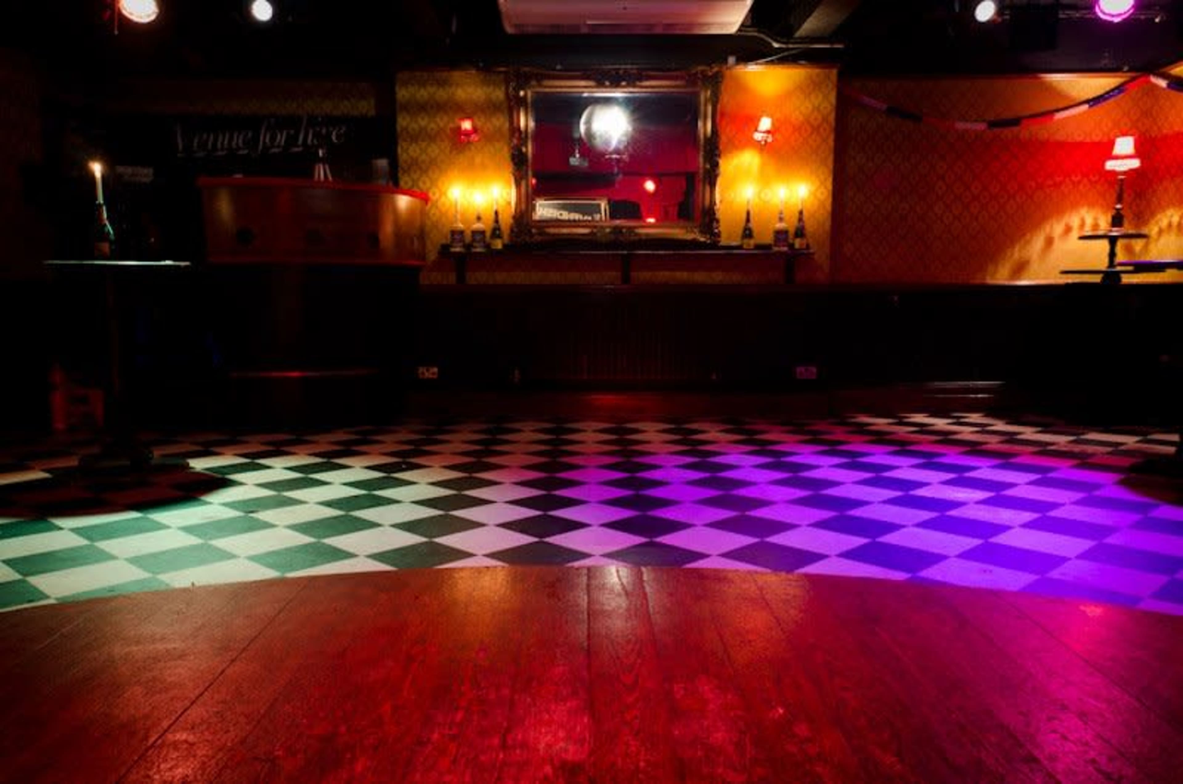 A dimly lit dance floor with a checkered black-and-white and wooden surface, surrounded by tables and lights.