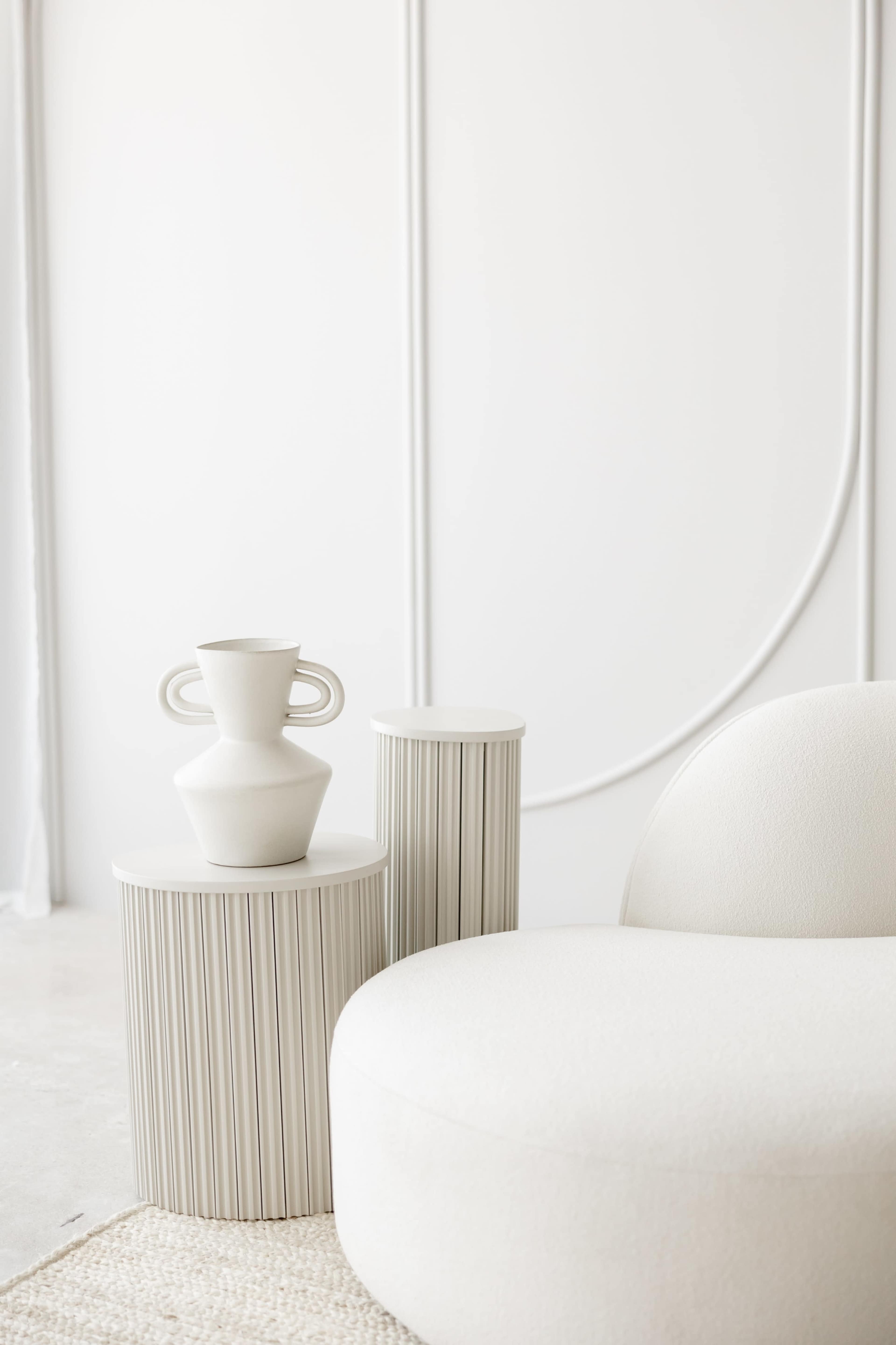 The image features a minimalist interior scene with a curved white sofa and two cylindrical side tables, one adorned with a decorative vase.