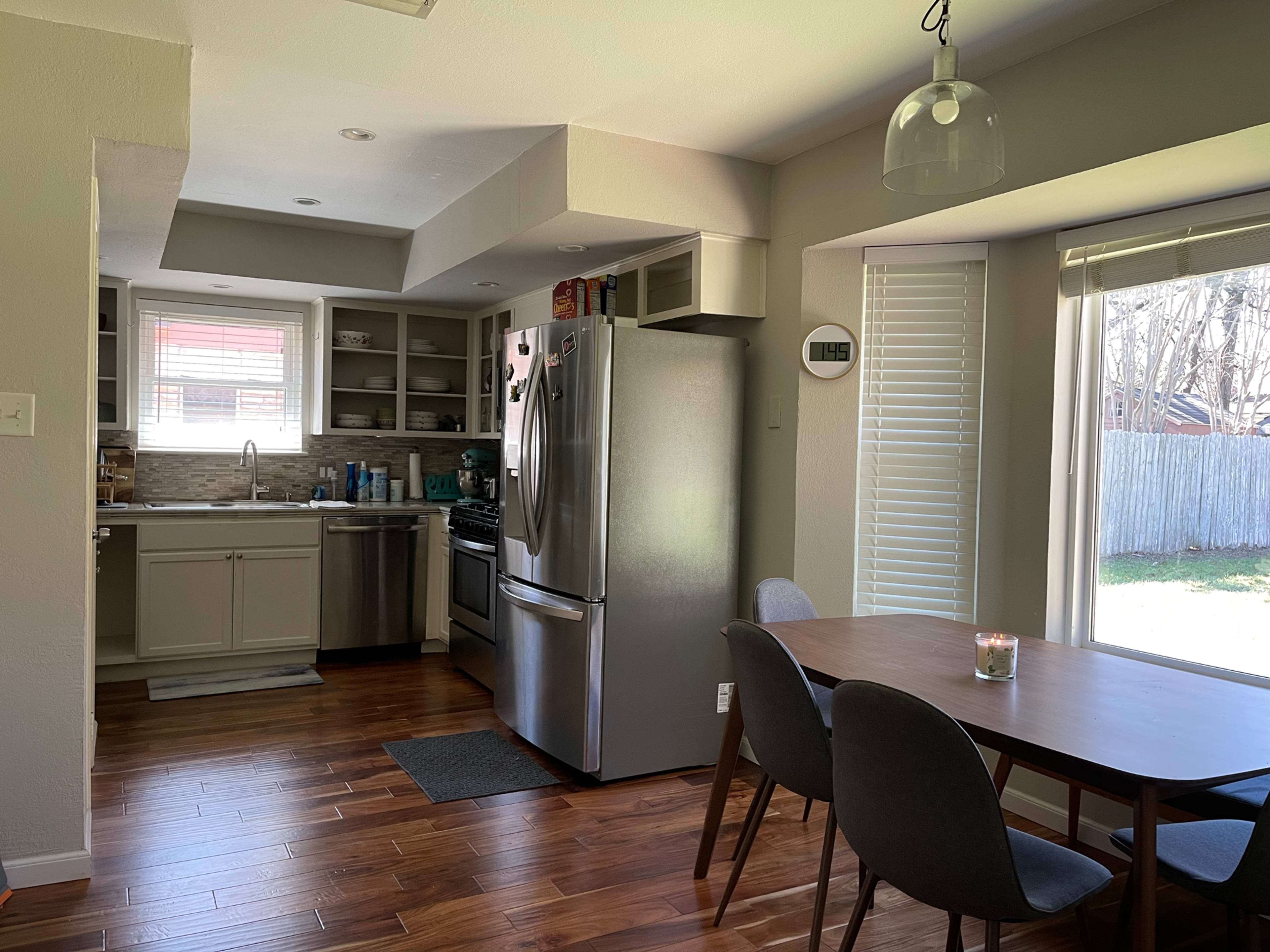 A modern kitchen with stainless steel appliances and a dining area featuring a wooden table and chairs, adjacent to a window overlooking a backyard.