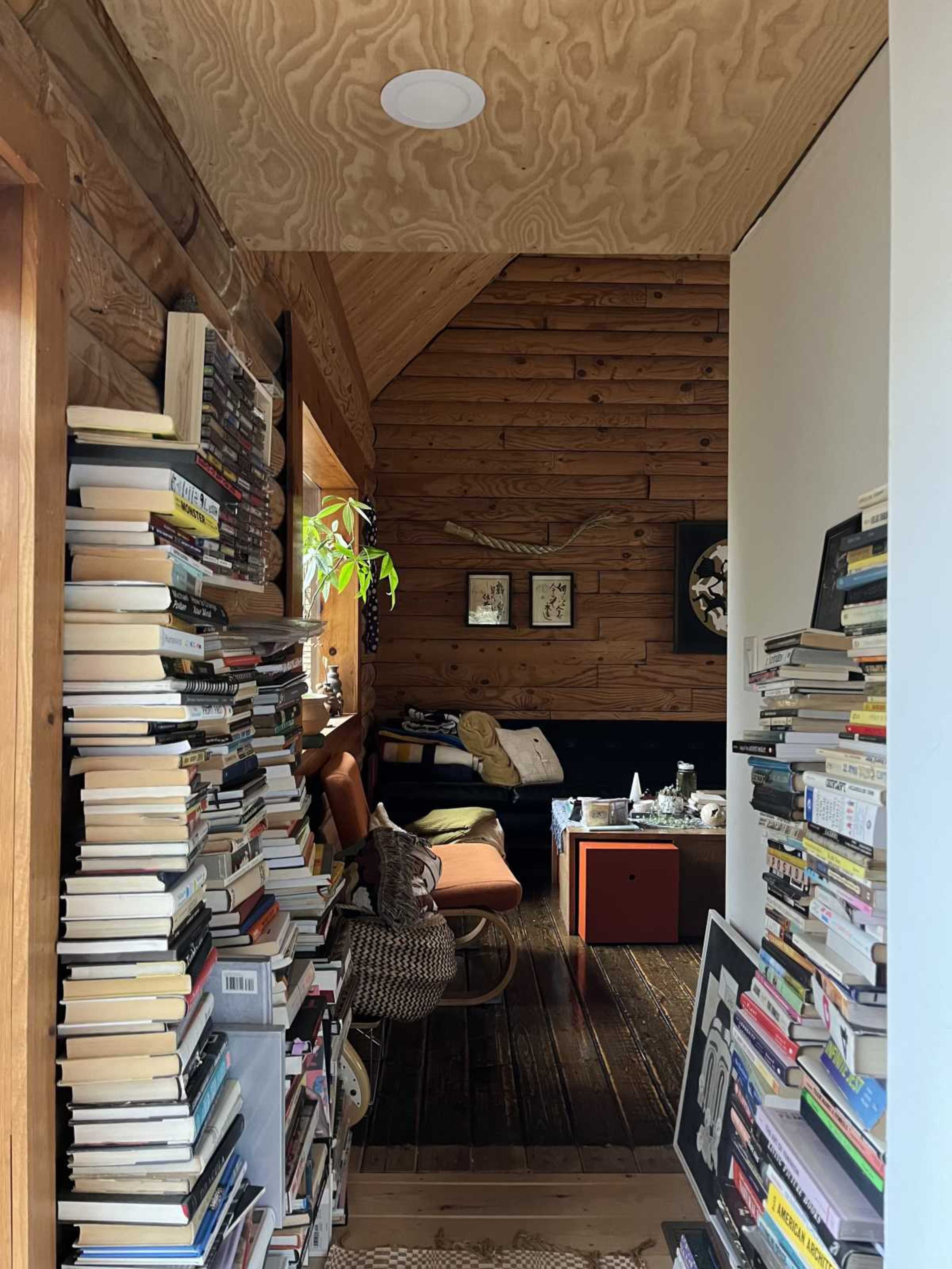 The image shows a narrow hallway lined with tall stacks of books on both sides, leading to a cozy living area at the back with wooden walls and furniture.