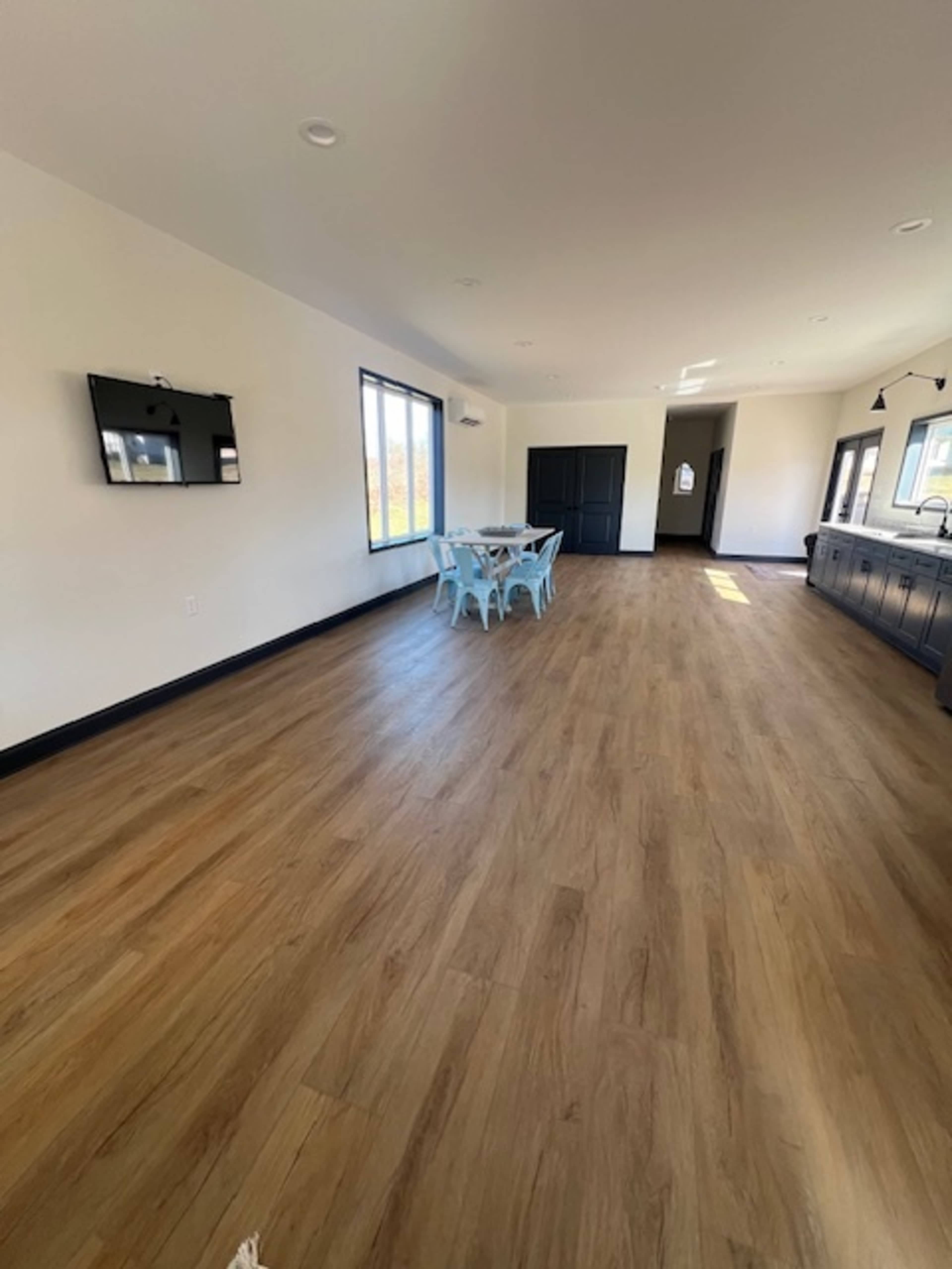 A spacious room features wooden flooring, large windows, and a dining area with a blue table and chairs.