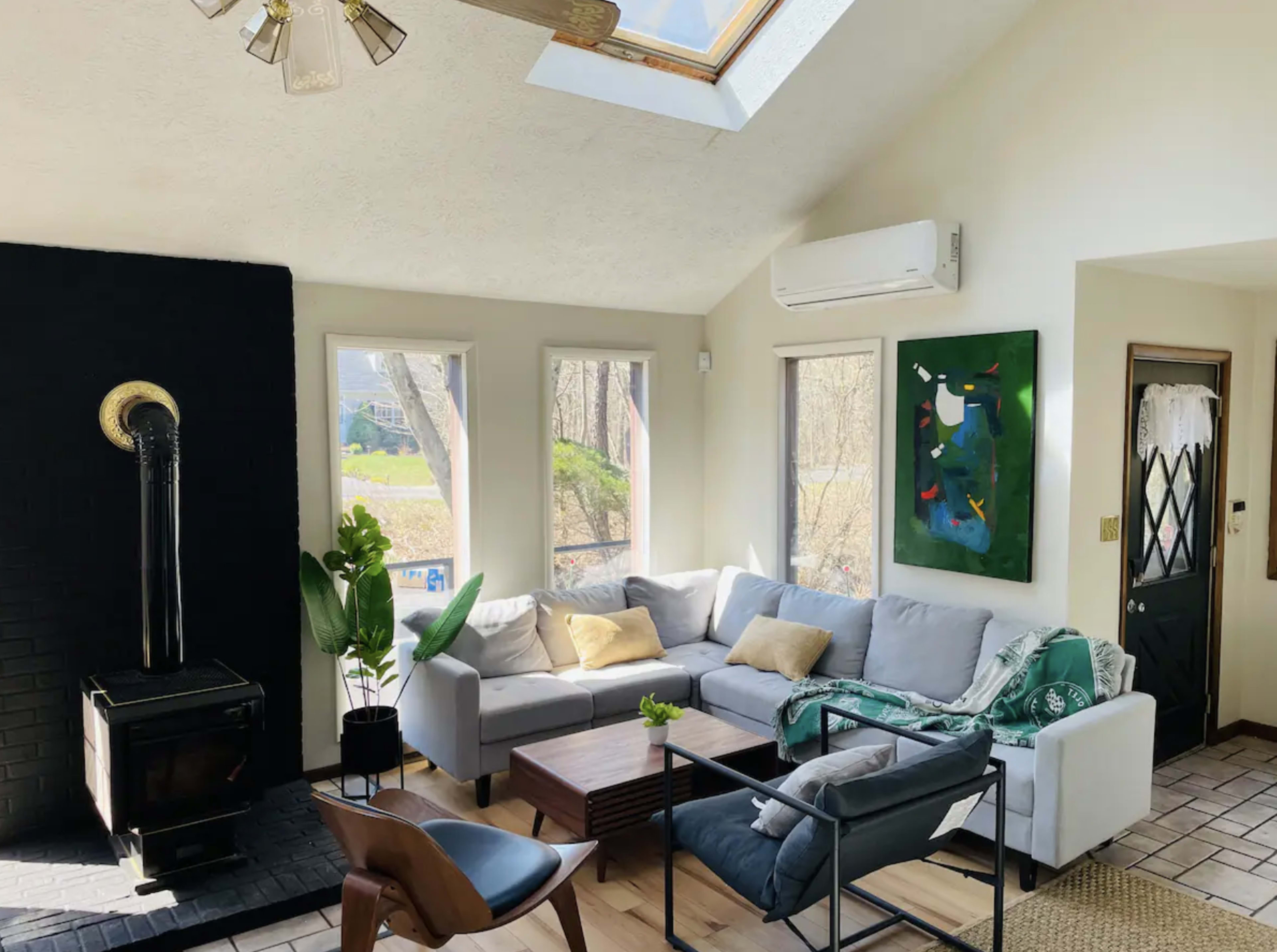 A bright living room features a sectional sofa, a coffee table, and large windows with a view, complemented by a wood-burning stove and a wall-mounted air conditioning unit.
