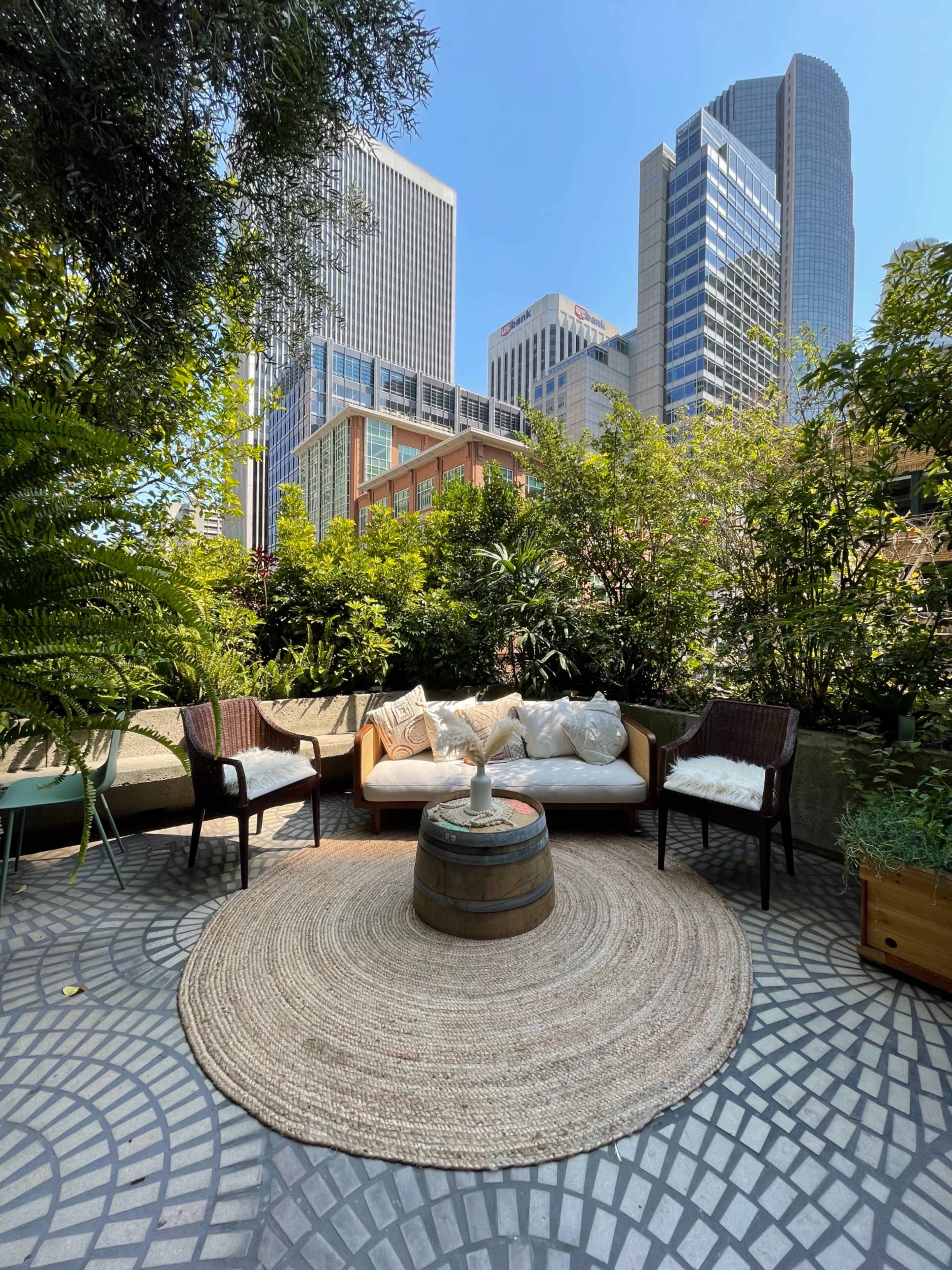 A cozy outdoor seating area featuring a circular rug and a fire pit, surrounded by greenery and tall buildings in the background.