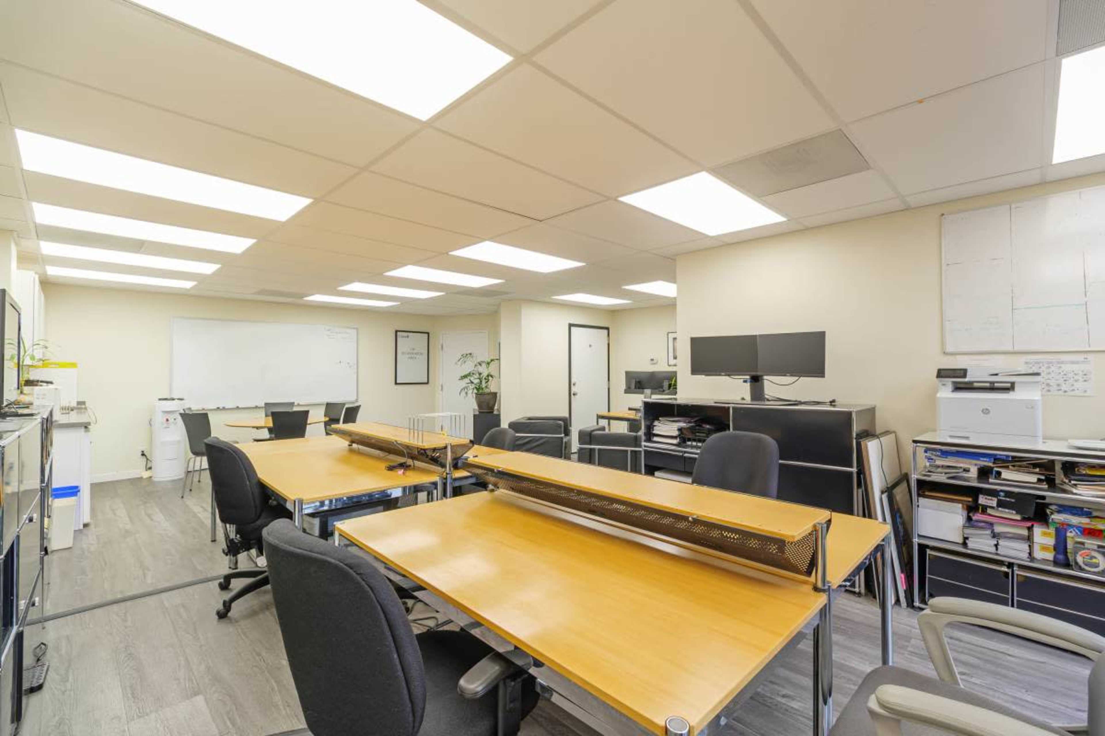 The image depicts a spacious office layout with several wooden desks, ergonomic chairs, and various office equipment, including monitors and filing cabinets.