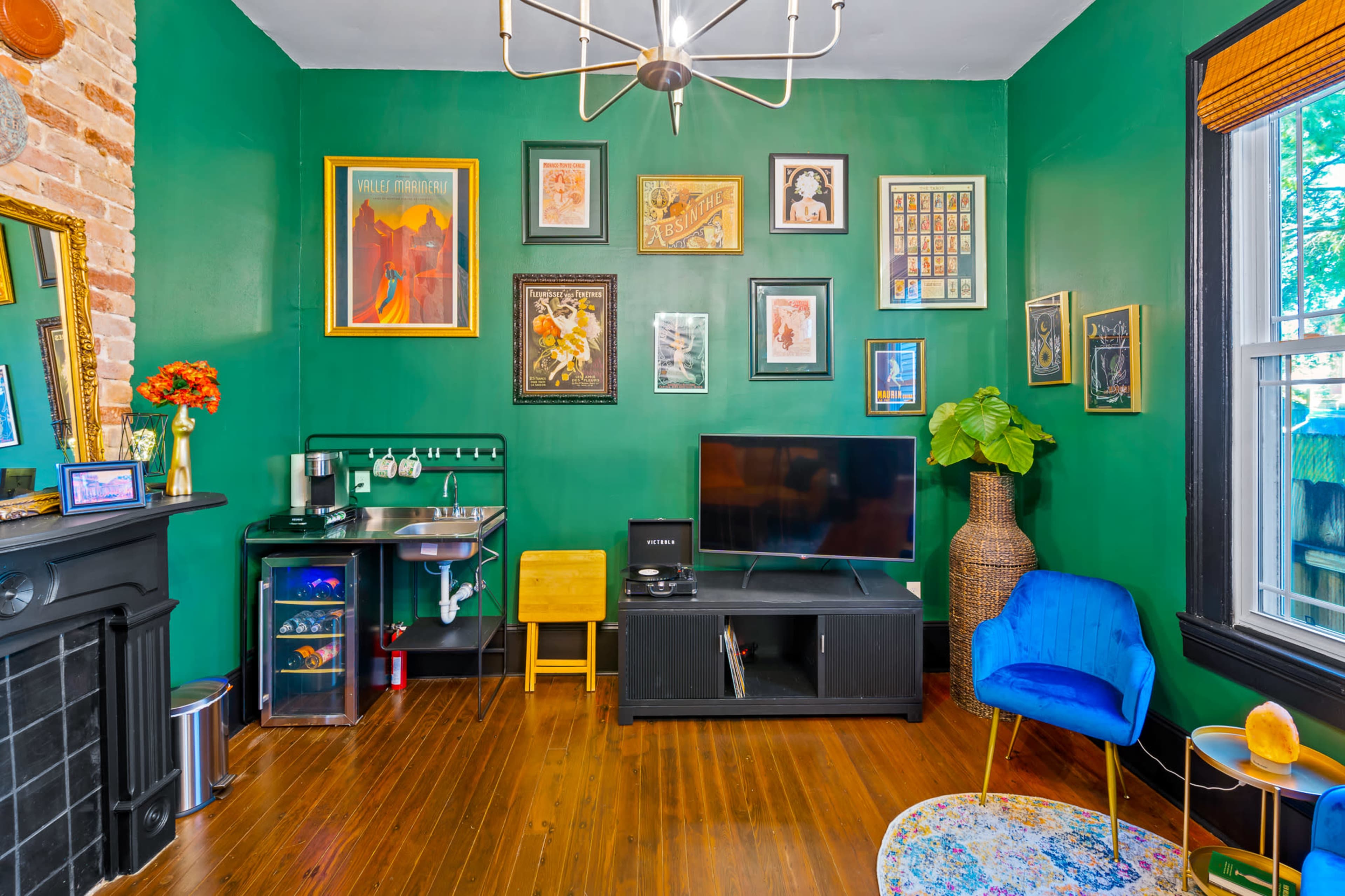 The scene features a room with green walls adorned with various framed artworks, a black TV stand with a television, a small kitchenette area, and a blue chair beside a potted plant.