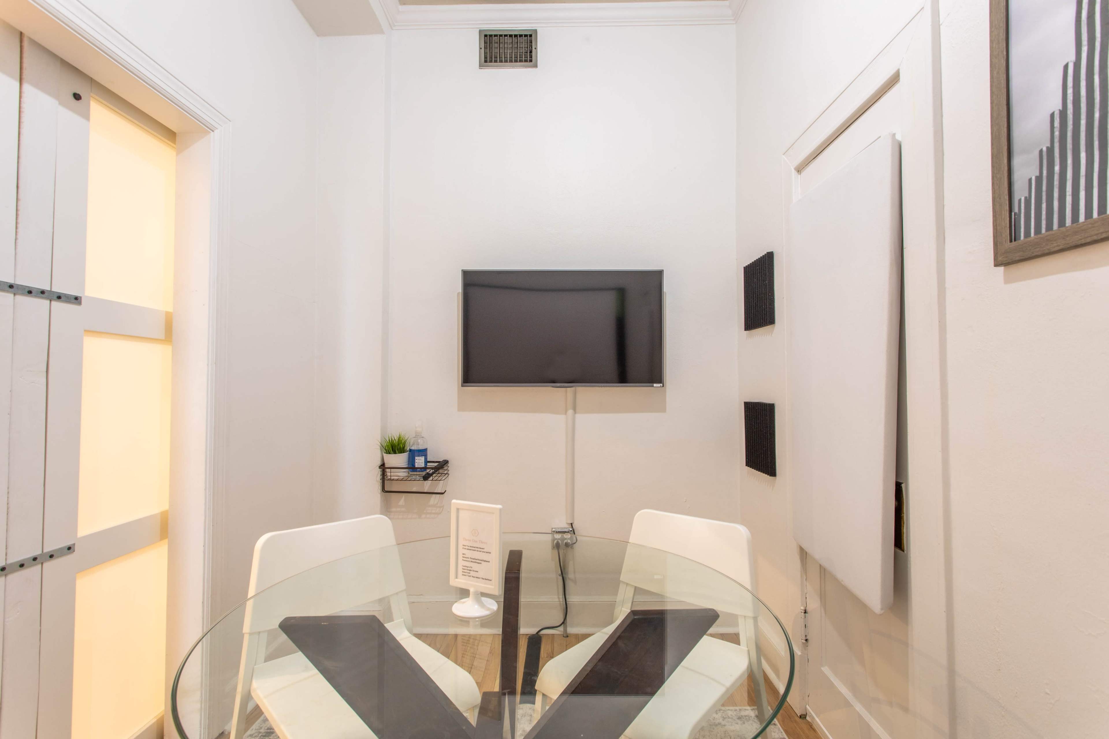 The image shows a small meeting room featuring a glass table, two white chairs, a wall-mounted TV, and a plant on a shelf.