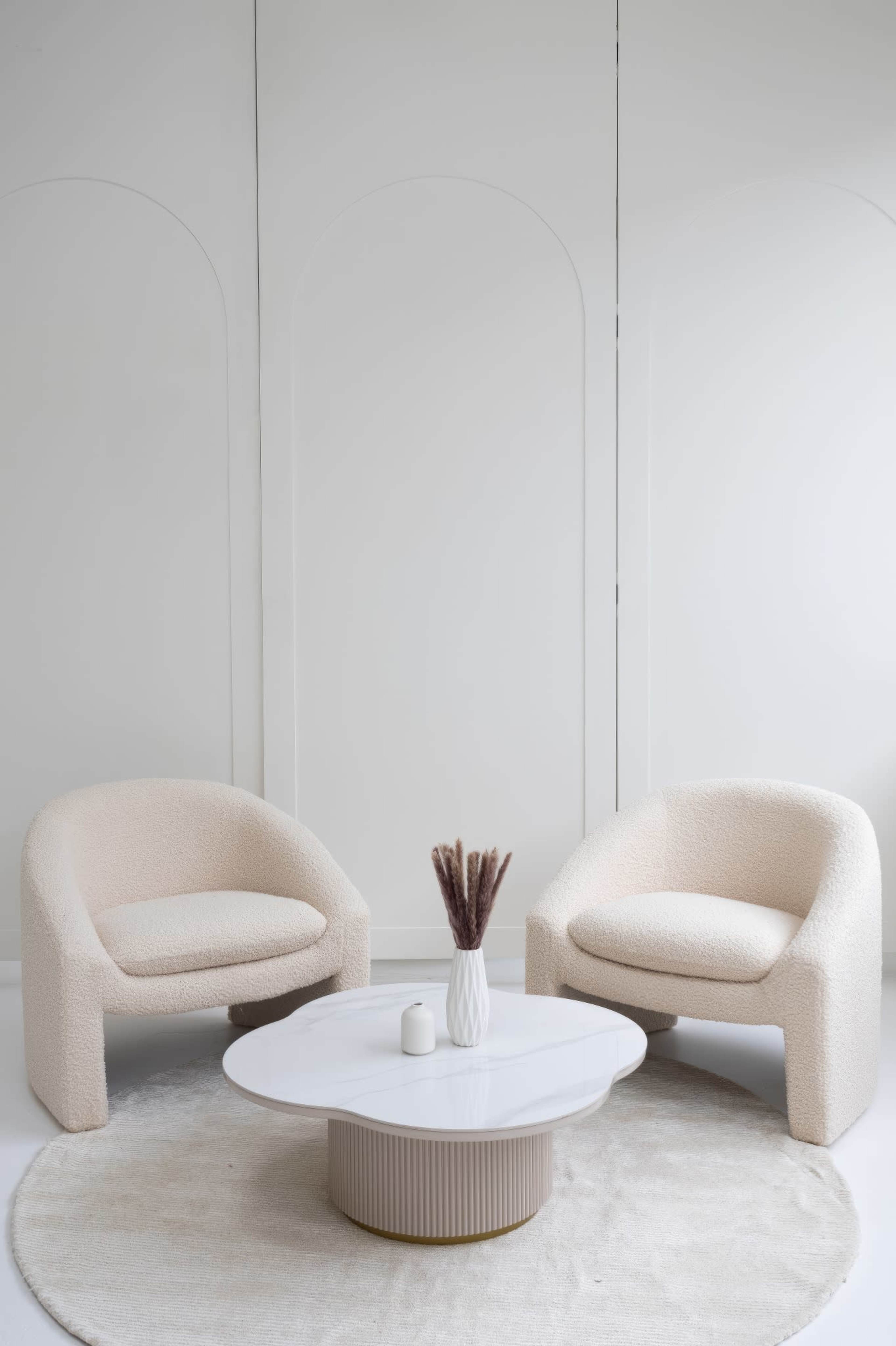 A pair of cream-colored armchairs are positioned on a circular beige rug facing a round marble coffee table, with a vase of dried flowers on top.