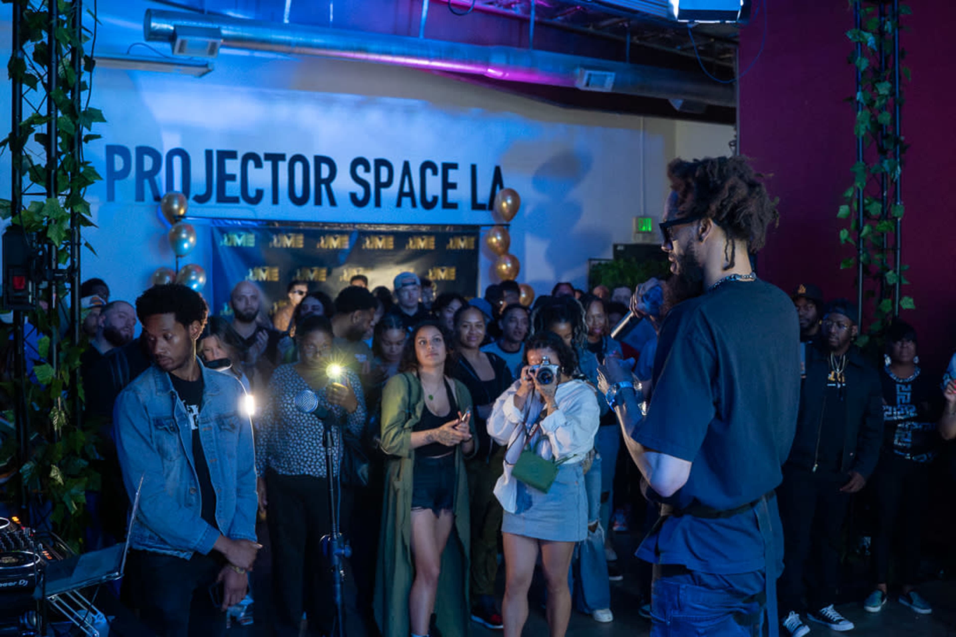 A crowd gathers in a venue named "Projector Space LA" as a speaker addresses them from the front.