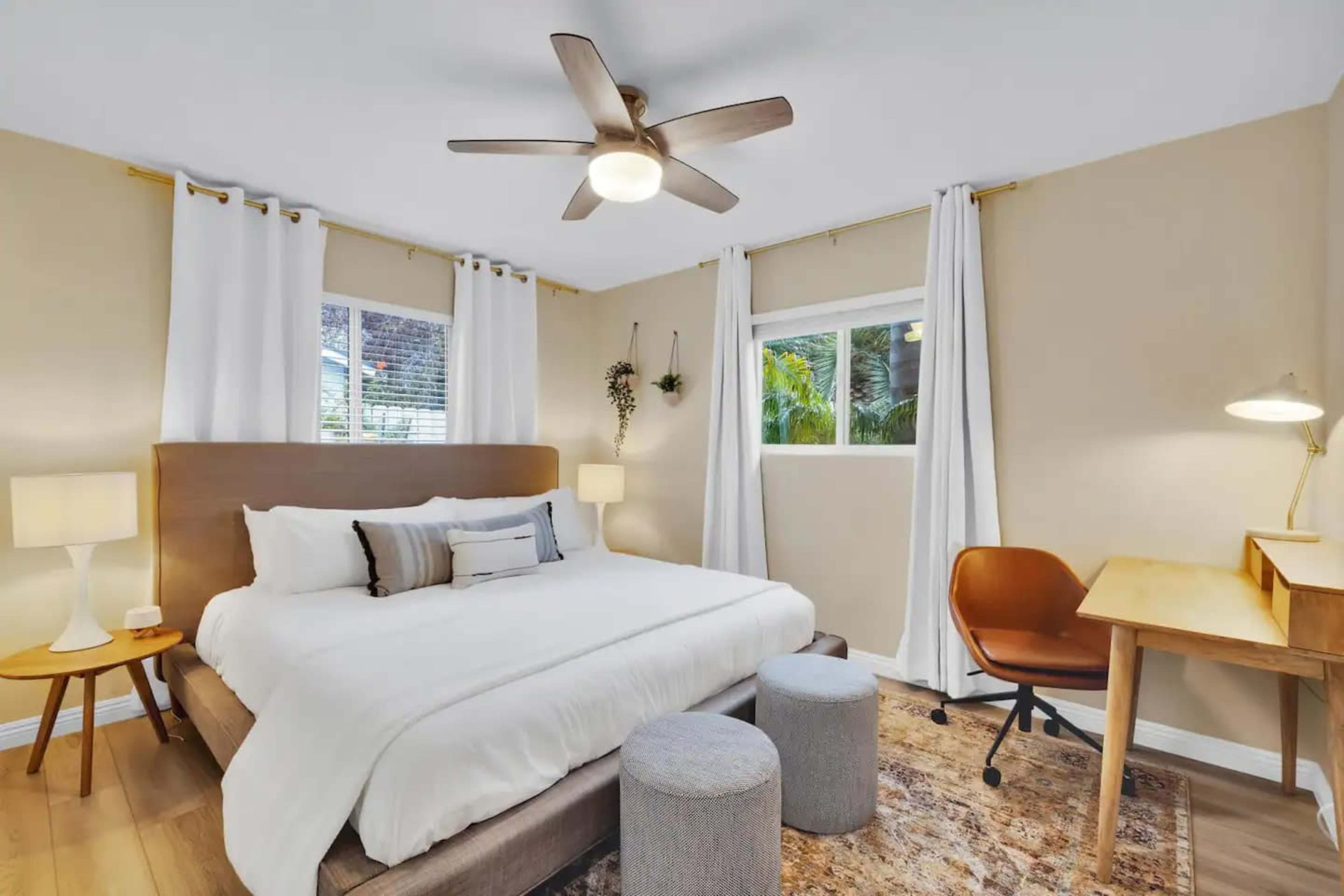 A modern bedroom features a large bed, two bedside lamps, a wooden desk with a chair, and large windows with white curtains.