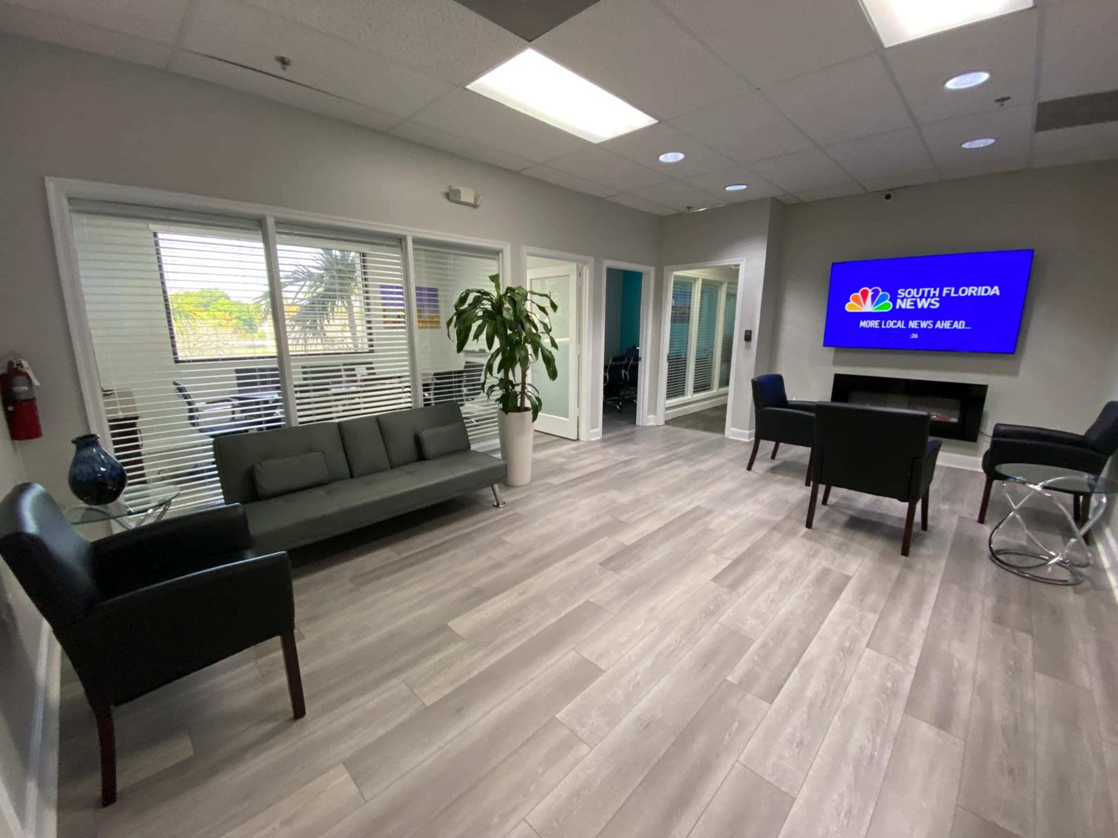 The image shows a modern waiting area with a gray couch, black chairs, a large television displaying a news channel, and a potted plant.