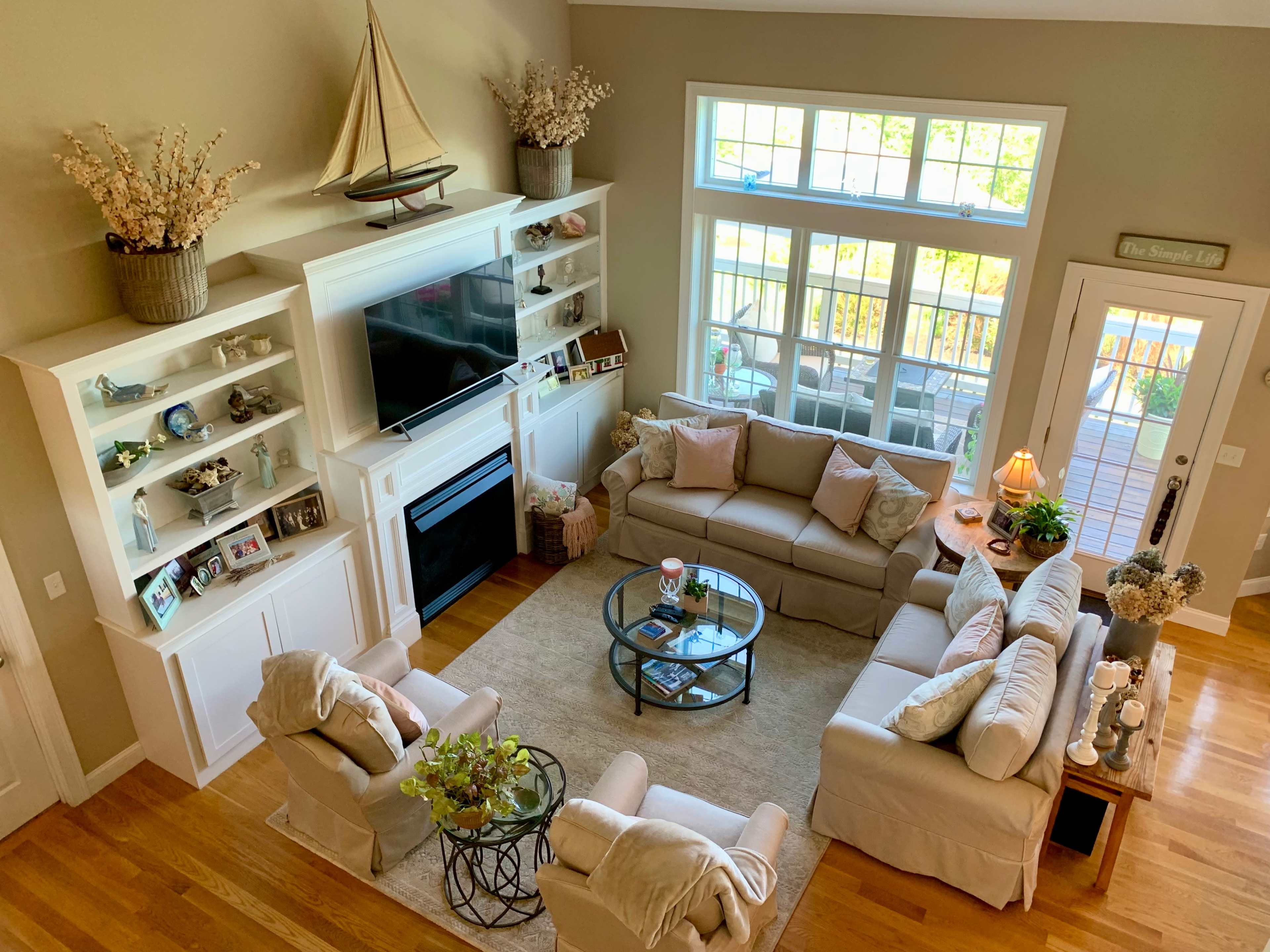 A well-lit living room features a central seating arrangement with a glass coffee table, surrounded by light-colored sofas and large windows showcasing a view outside.