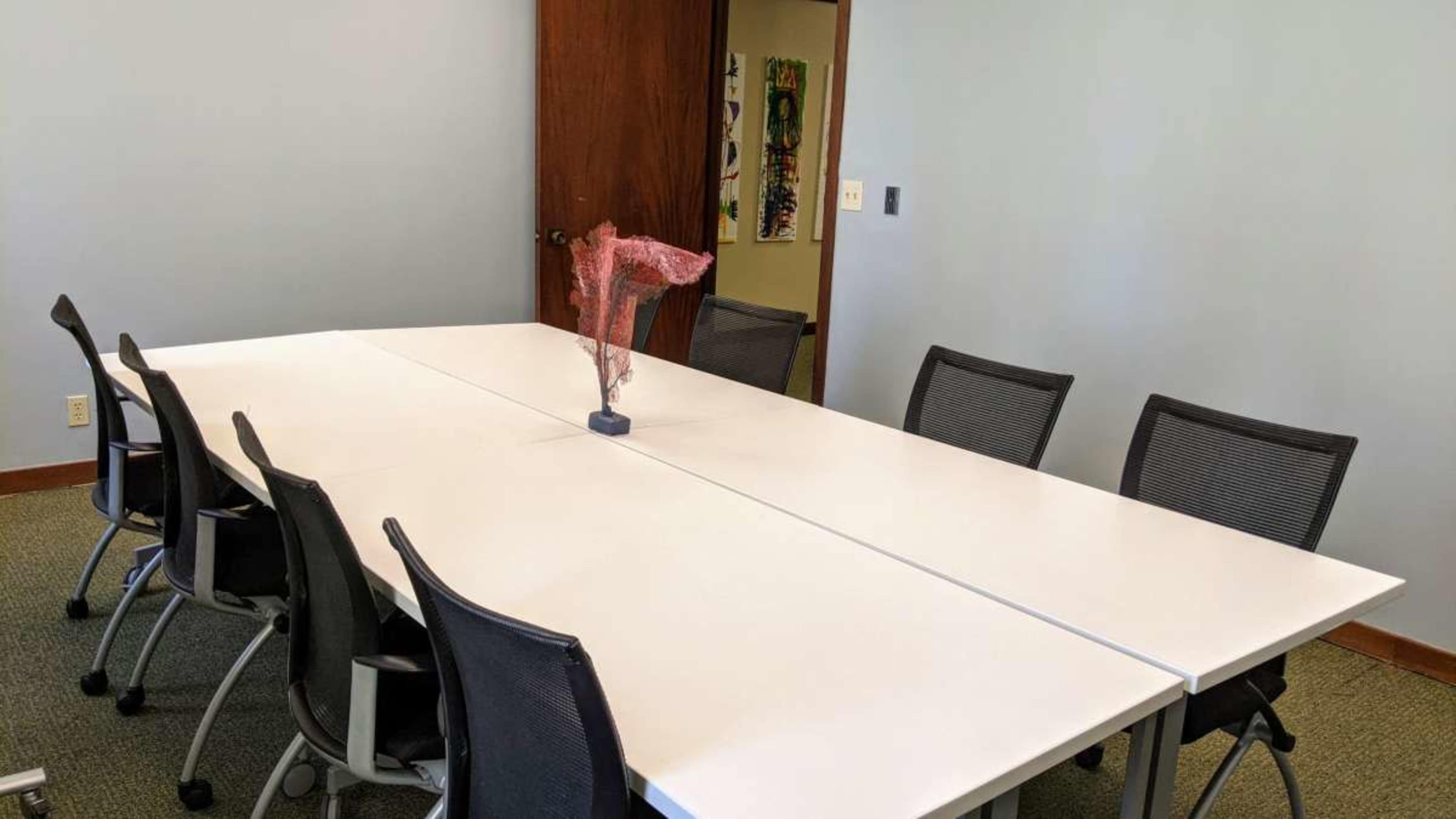 A modern conference room features a long white table with a decorative piece at the center and several black office chairs arranged around it.