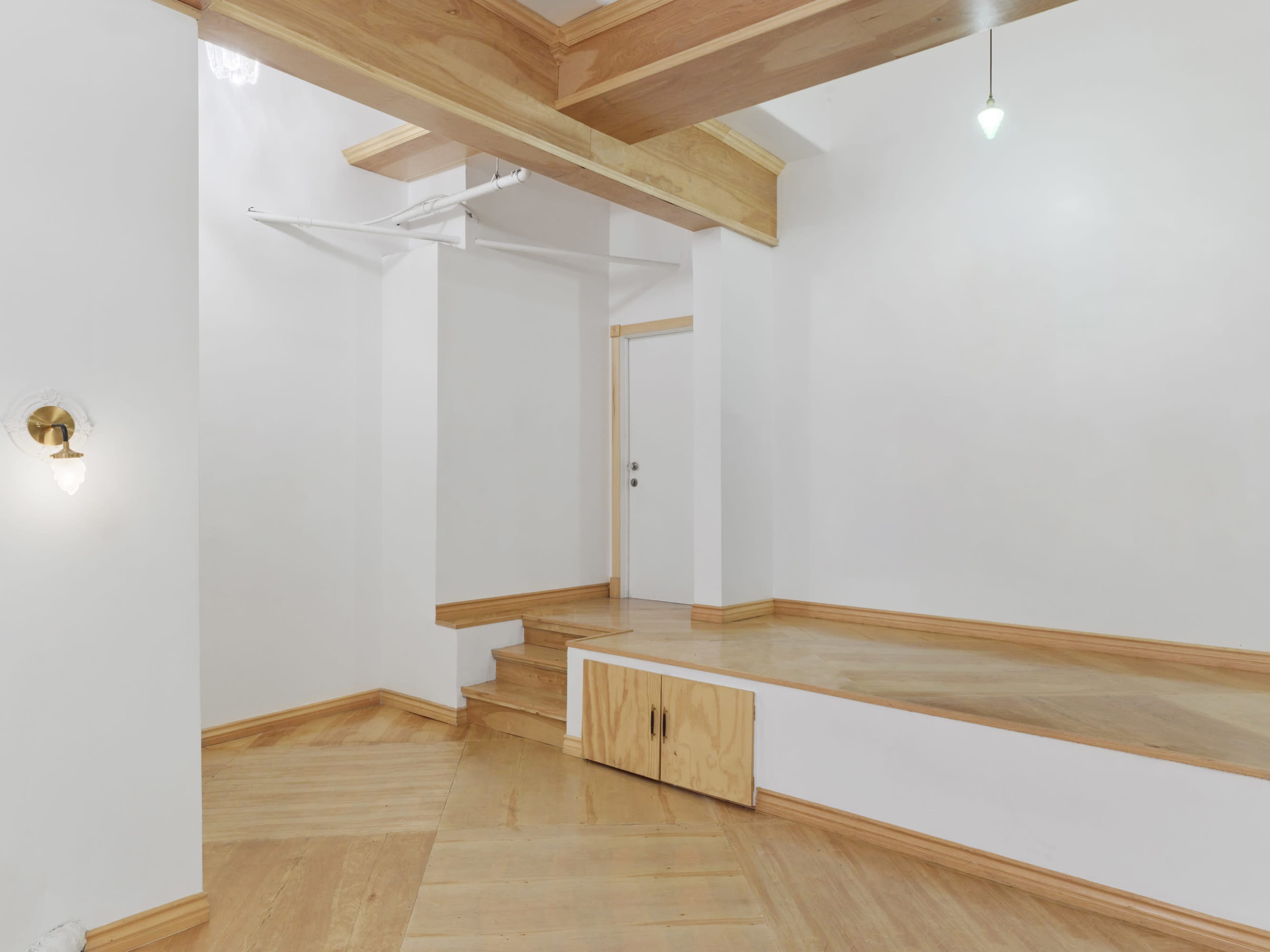 The image shows a minimalist room with wooden flooring, a small set of stairs, and a closed door at the far end.