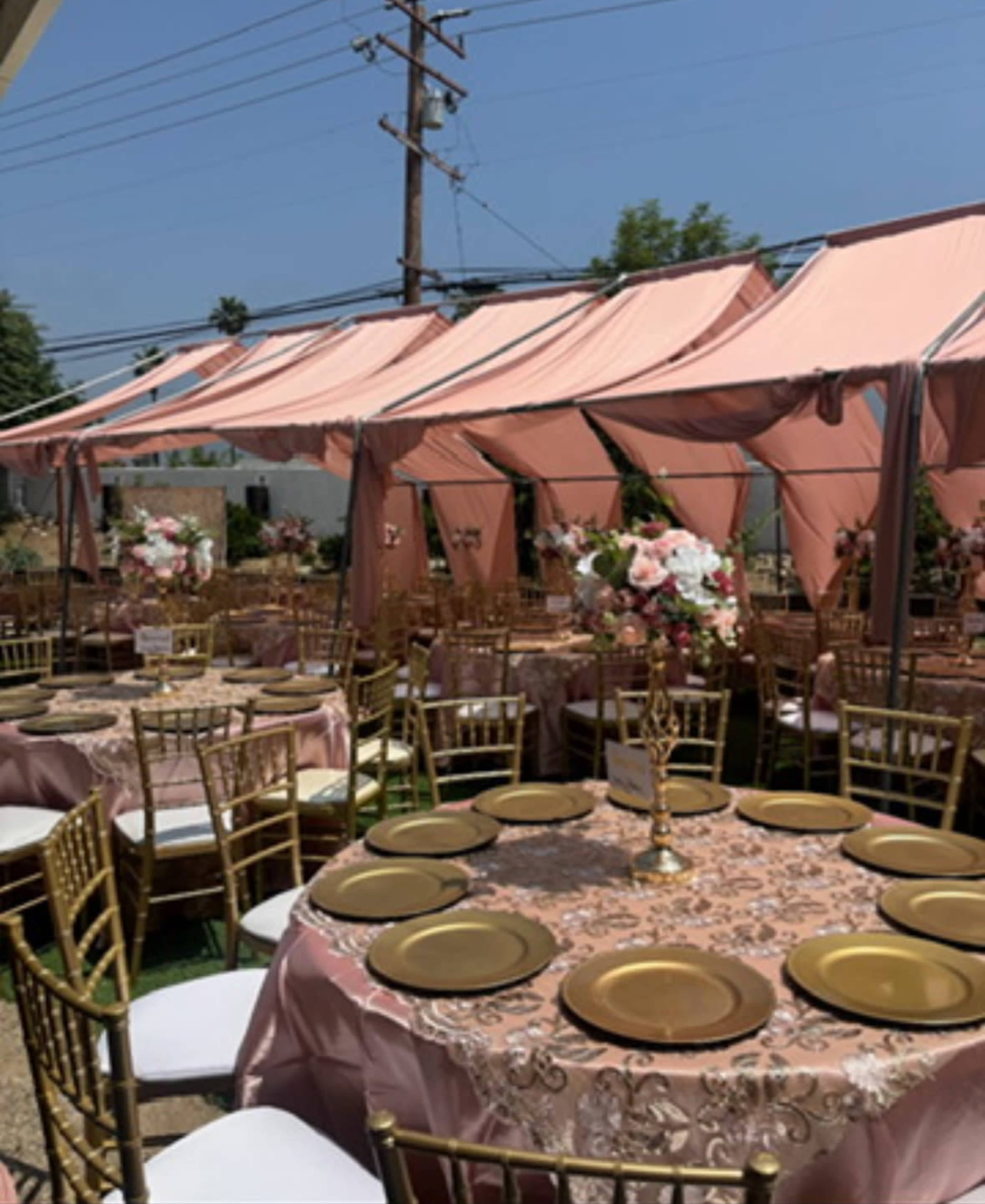 The image shows a decorated outdoor event space with round tables set with gold plates and floral centerpieces under a pink tent.