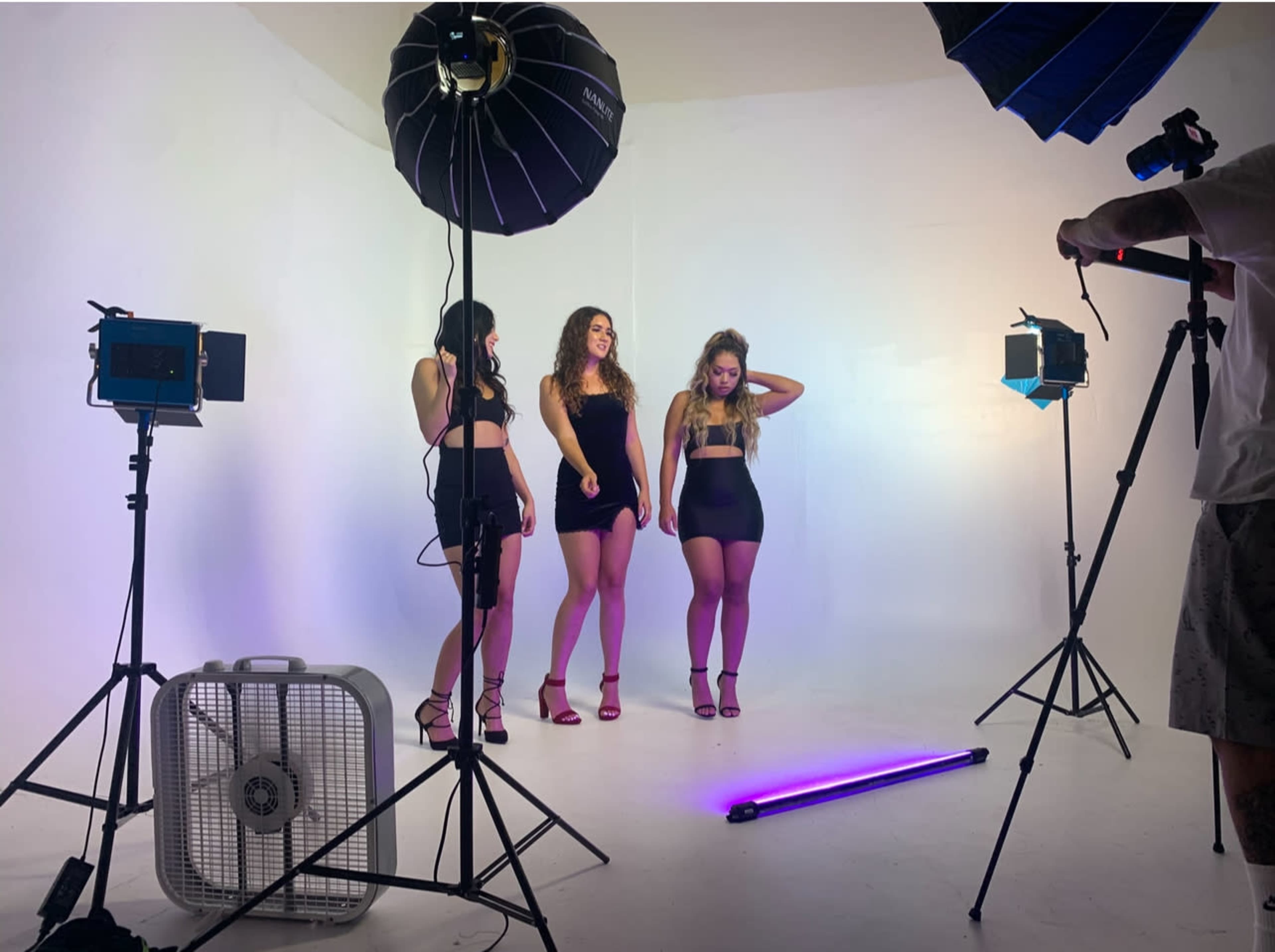 Three models pose on a white backdrop while a photographer captures the scene using studio lighting and equipment.