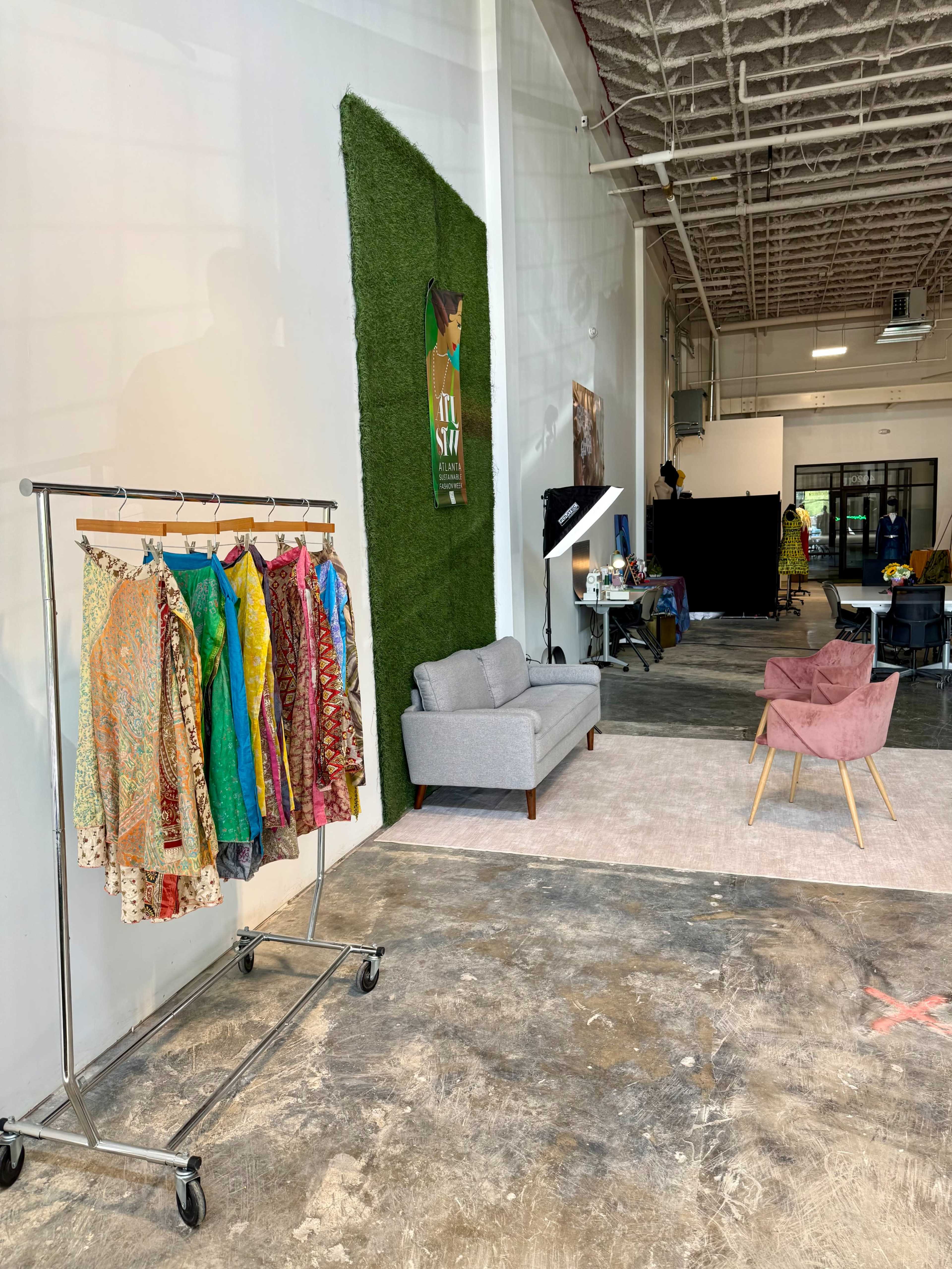 The image shows a spacious interior with a clothing rack displaying colorful garments, a gray sofa, and a pink armchair on a carpeted area against a backdrop of artificial grass and a wall with decor.