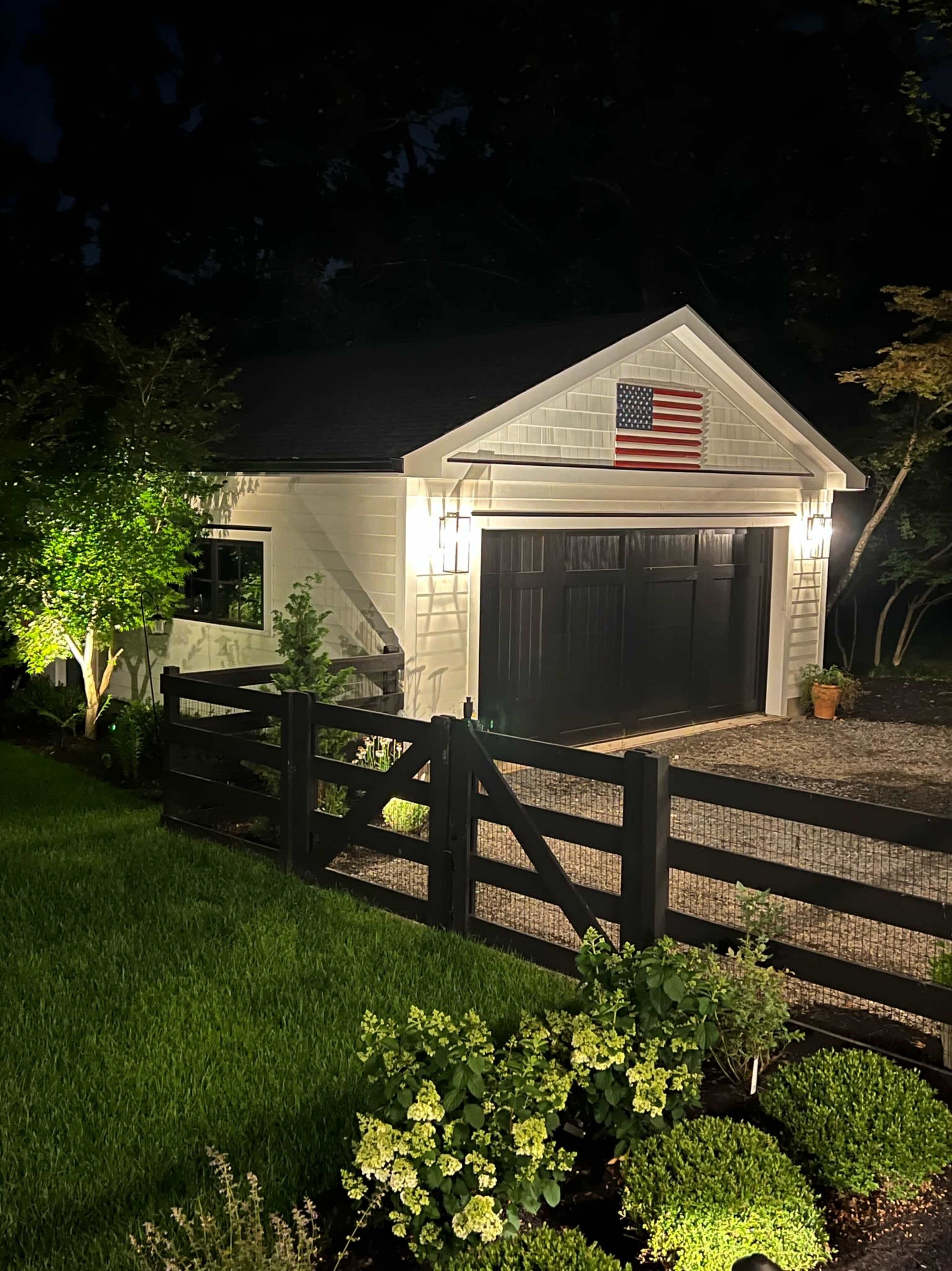 A two-car garage with a flag displayed on the front is illuminated at night, surrounded by landscaped greenery and a wooden fence.