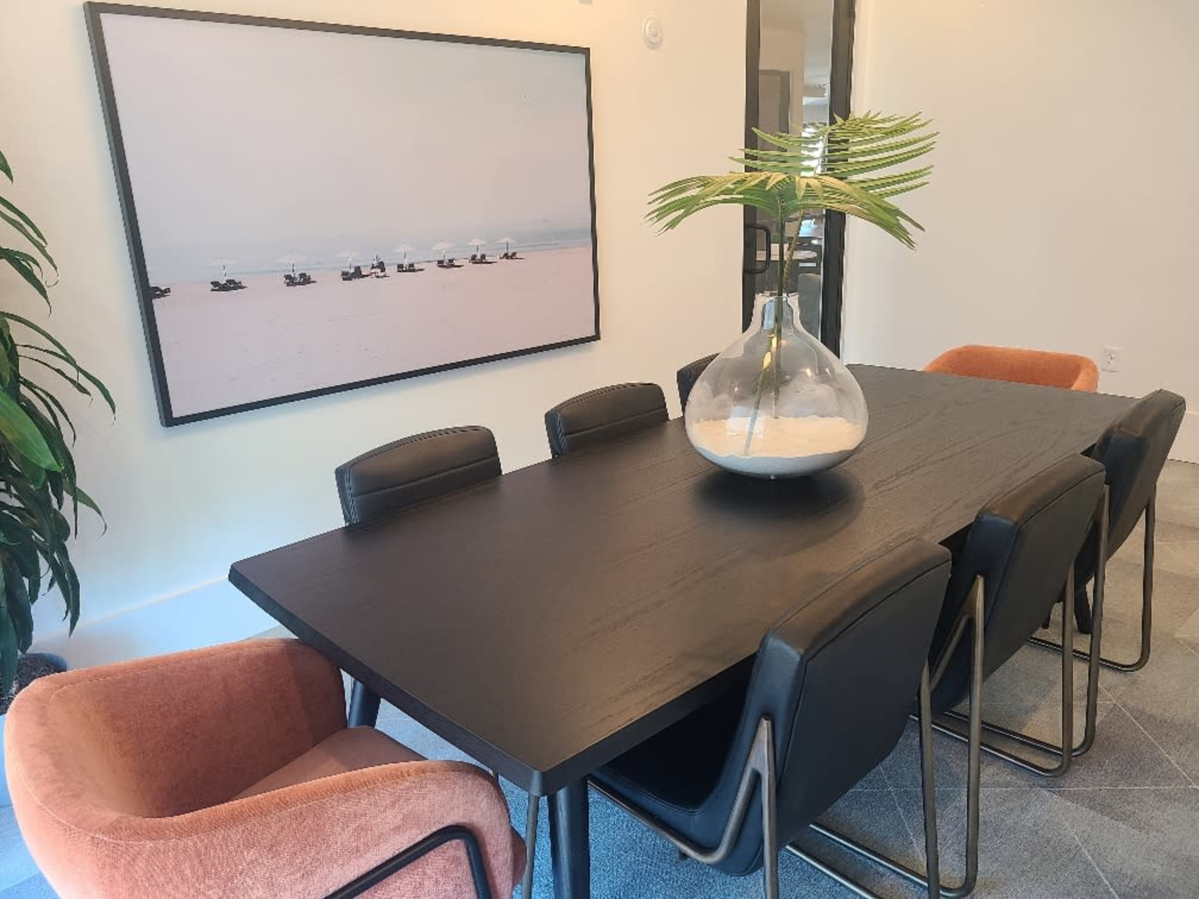 A dining table with black chairs and an orange accent chair is set against a large photograph of boats on a beach, with a glass vase containing a plant as the centerpiece.