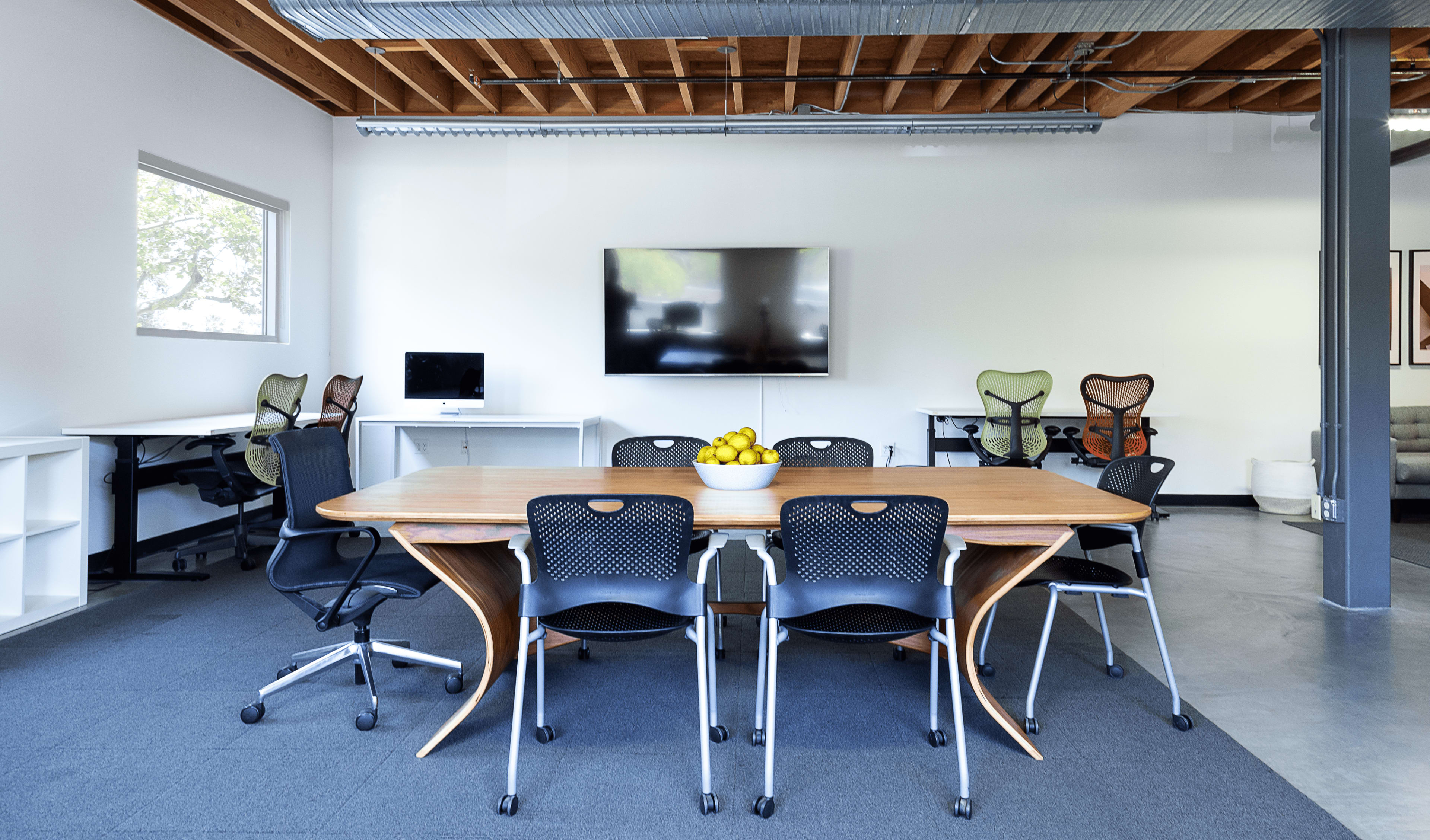 A modern office space featuring a wooden conference table surrounded by ergonomic chairs, with a large screen on the wall and workspace areas in the background.
