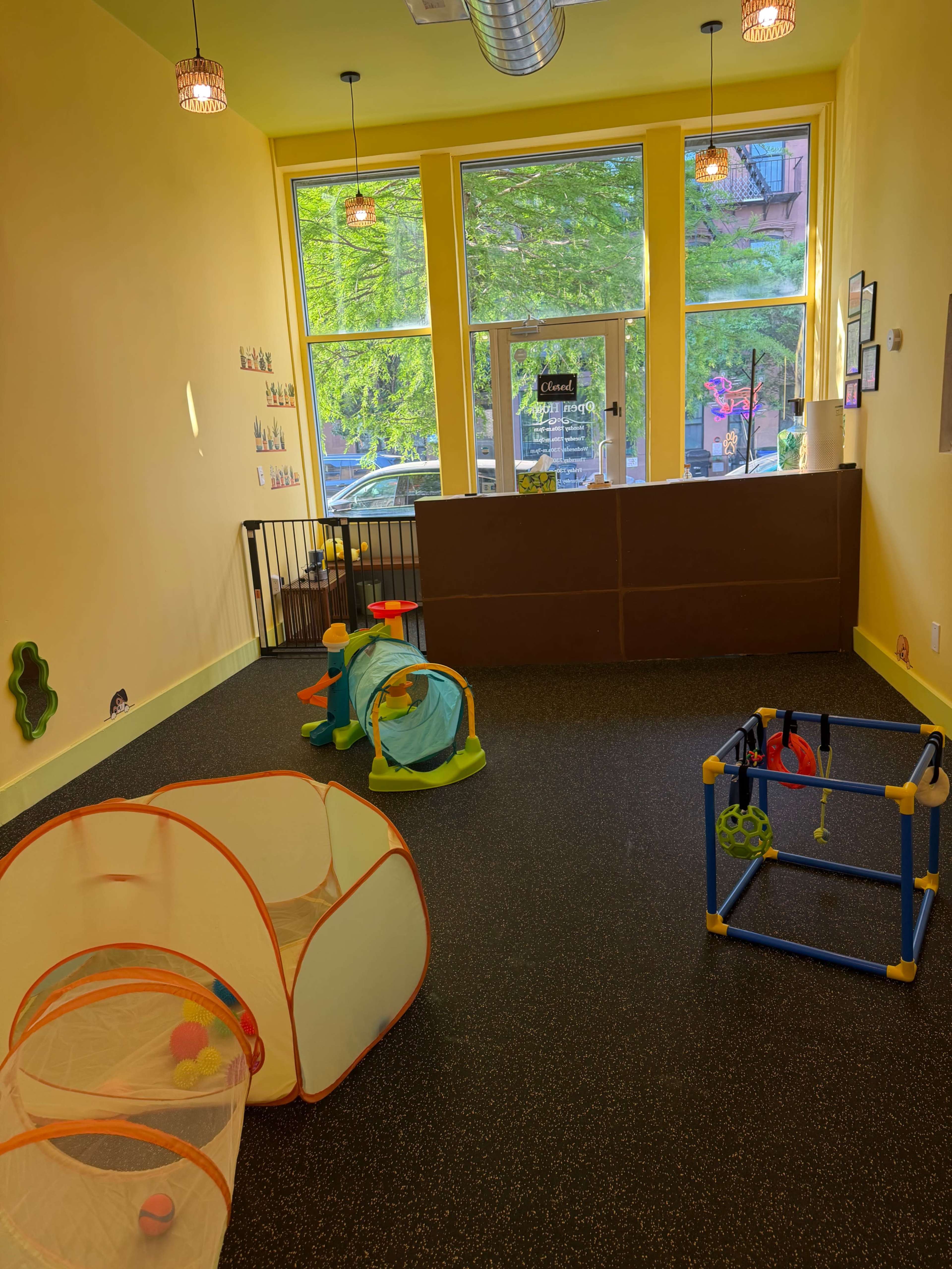 The image shows a brightly lit children's playroom with two colorful play tents, a ball pit, and a wooden activity center on a rubber floor.