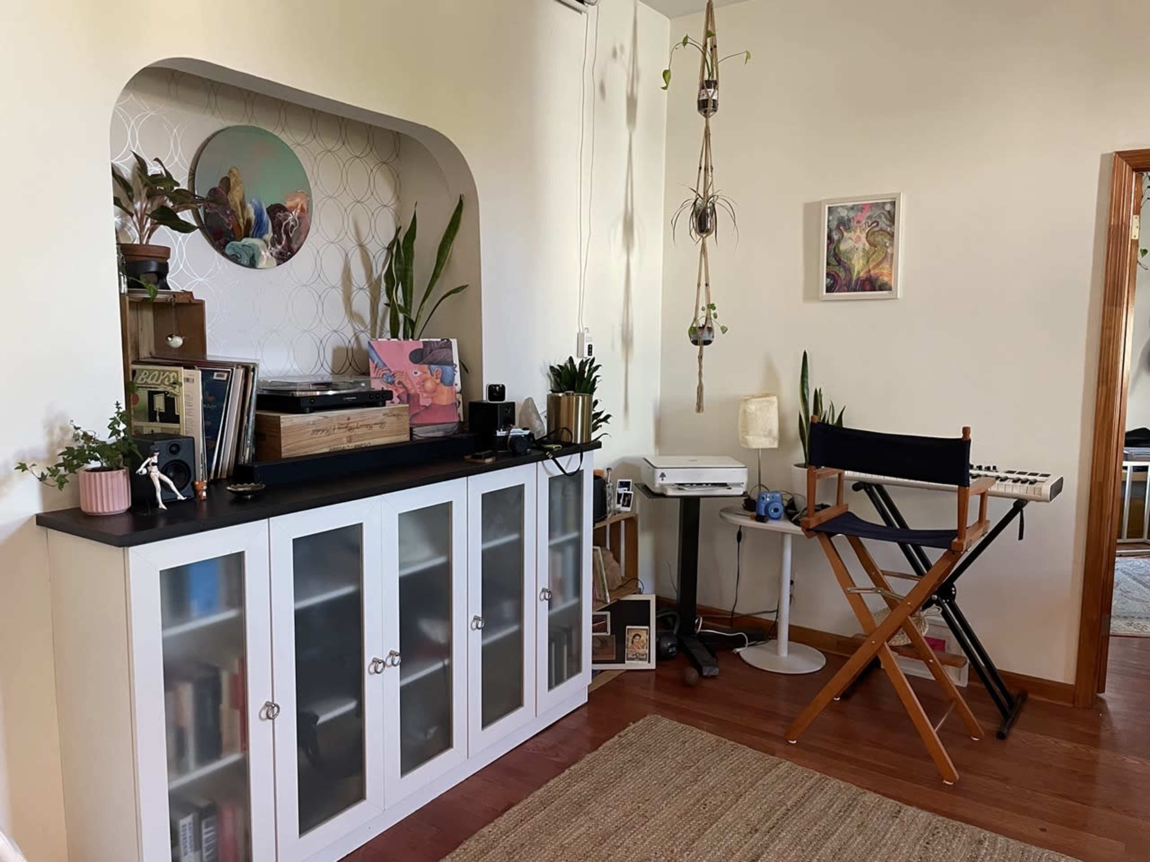 A cozy room featuring a white cabinet with glass doors, a record player, various plants, and a foldable director's chair alongside a small printer and keyboard.