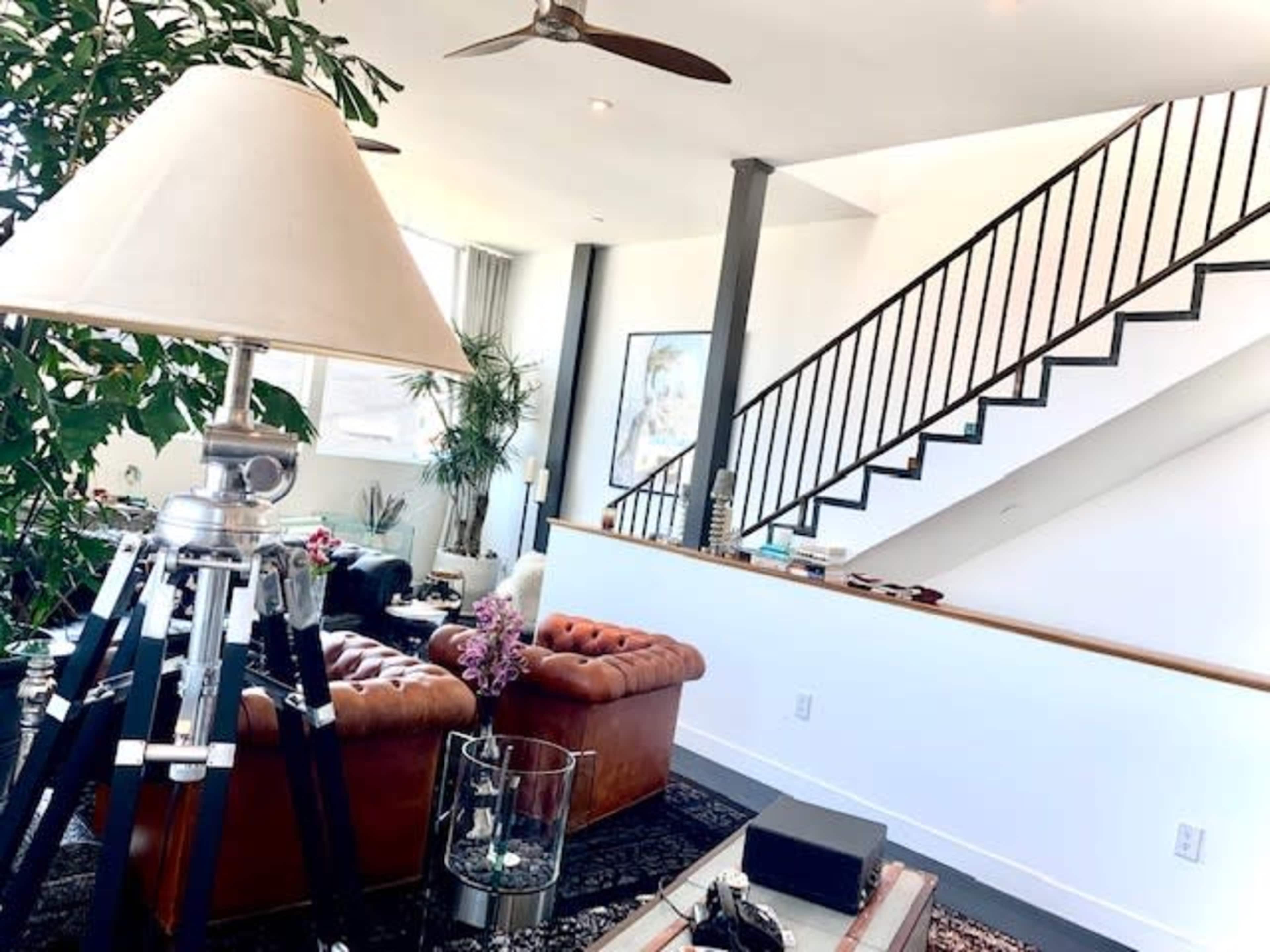 A modern living space with a staircase in the background, a leather sofa, a tripod lamp, and various decorative plants.