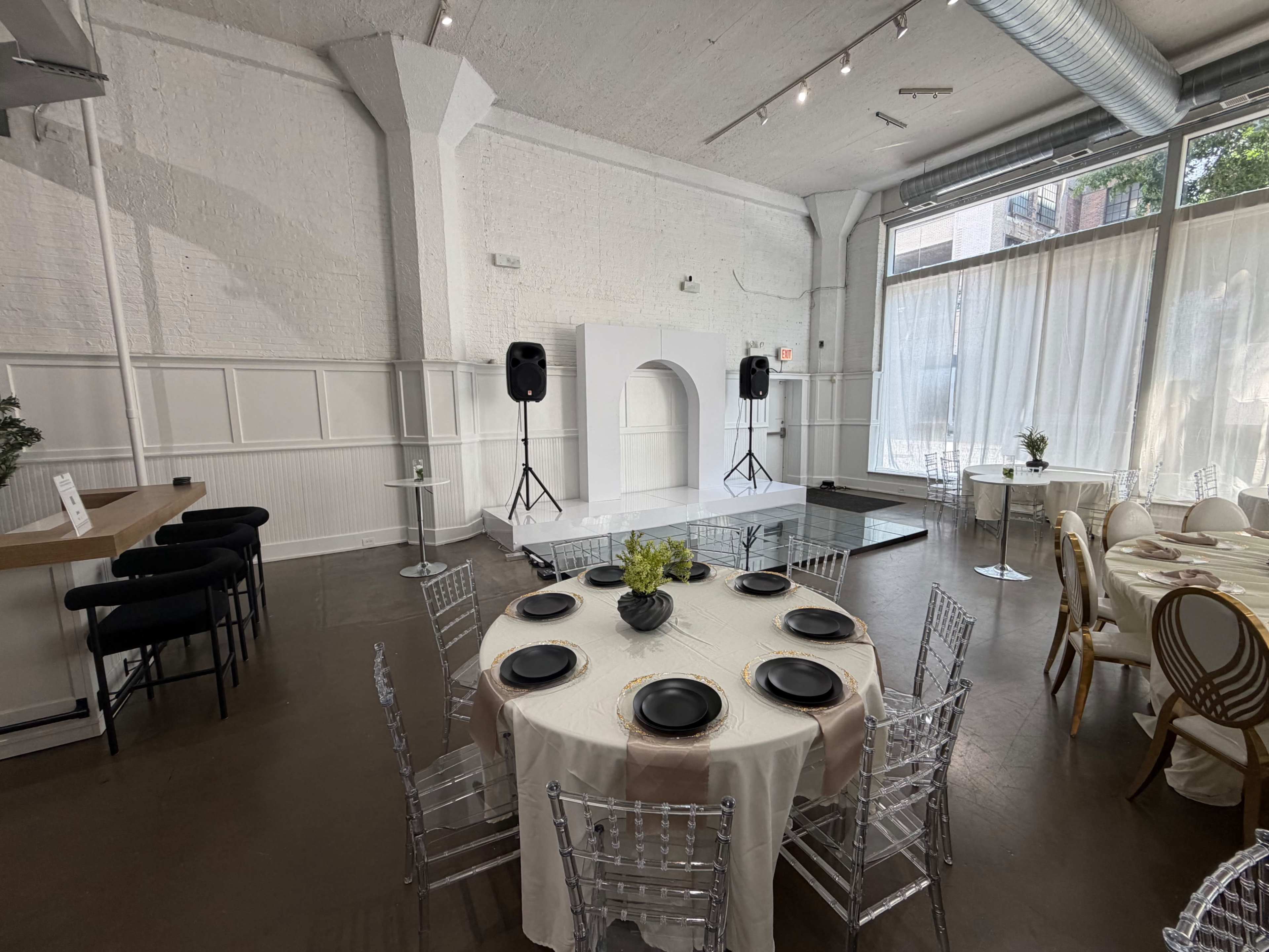 The image shows a spacious event venue featuring a circular table set for dining with transparent chairs, a simple stage with speakers, and large windows allowing natural light to enter.