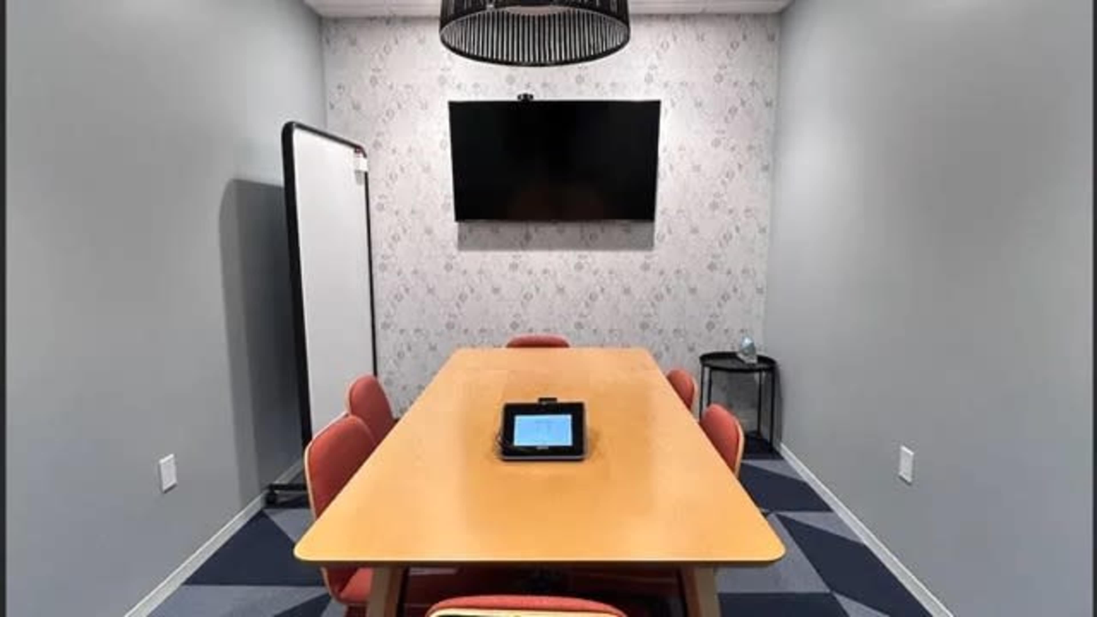 A conference room features a wooden table surrounded by five chairs, a television mounted on the wall, and a whiteboard nearby.