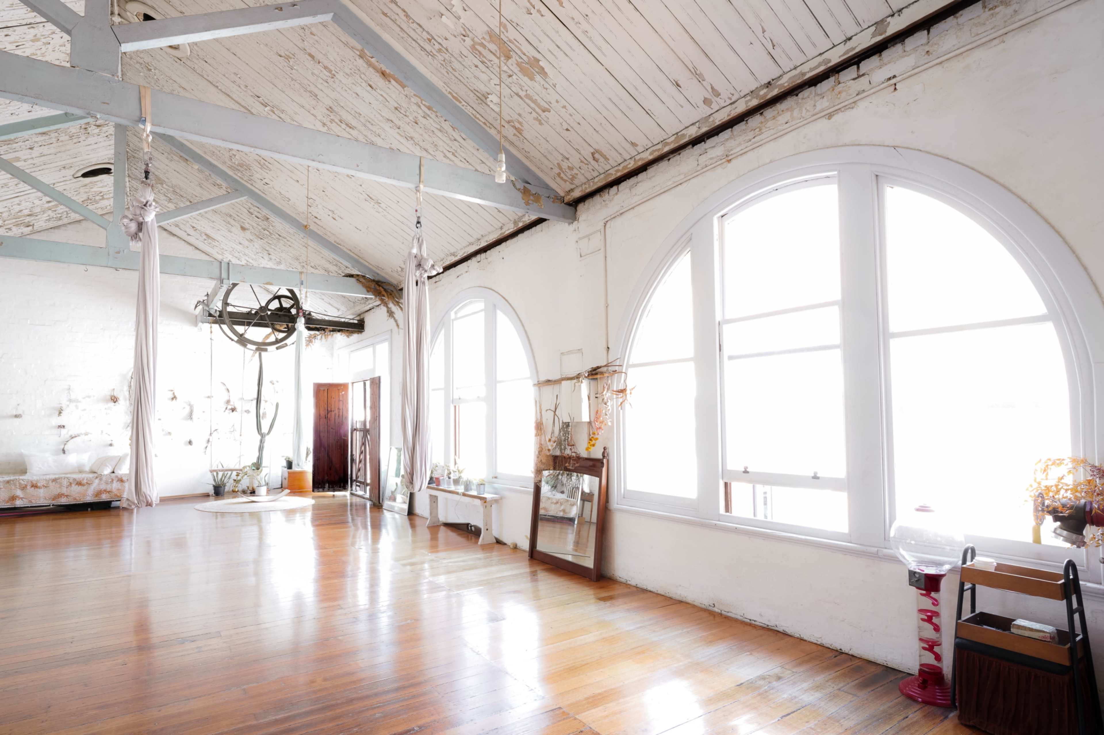 The image depicts a spacious, well-lit room with large arched windows, wooden floors, and exposed beams on the ceiling.