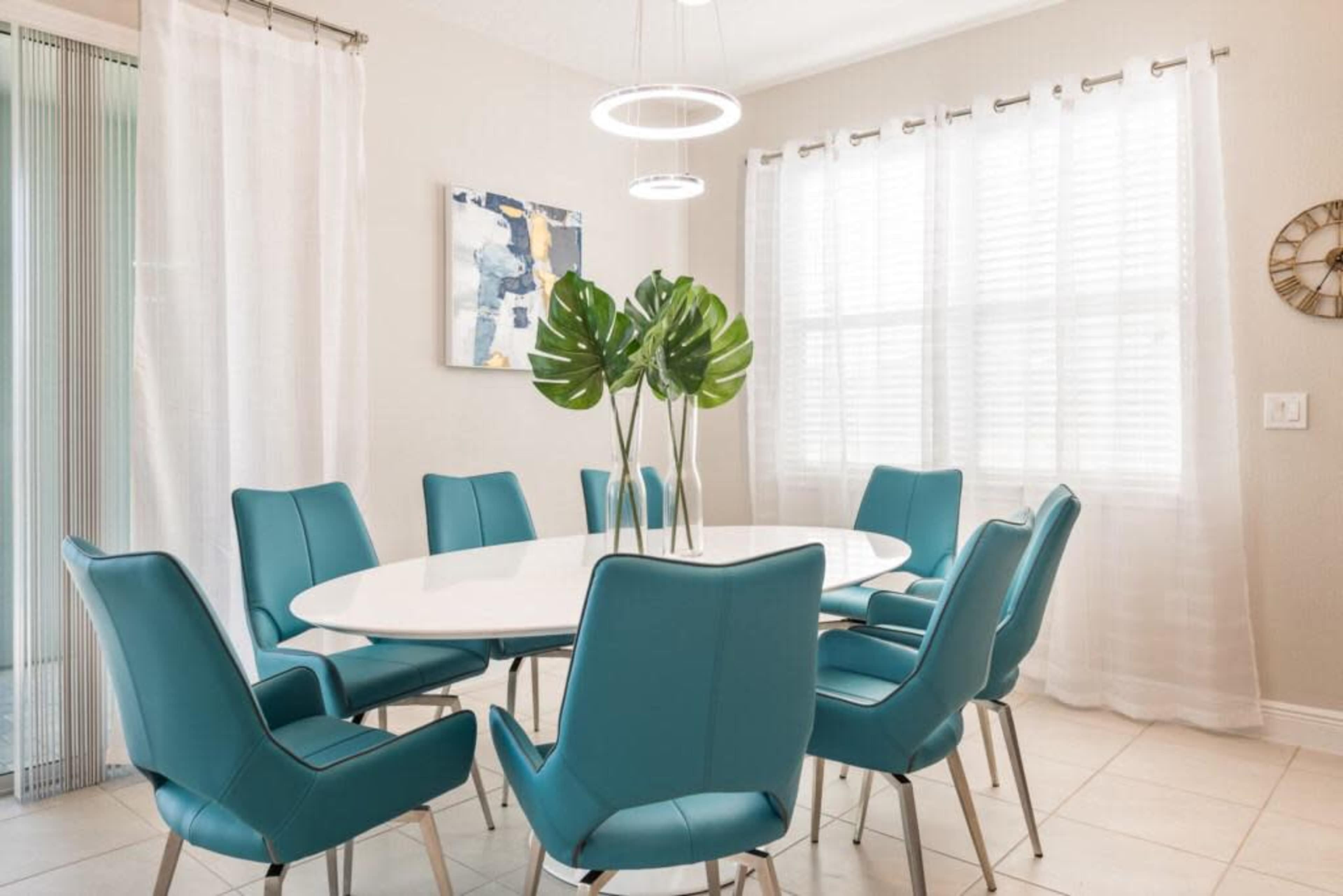 A modern dining area features an oval white table surrounded by eight teal chairs, with a decorative plant centerpiece and large windows letting in natural light.