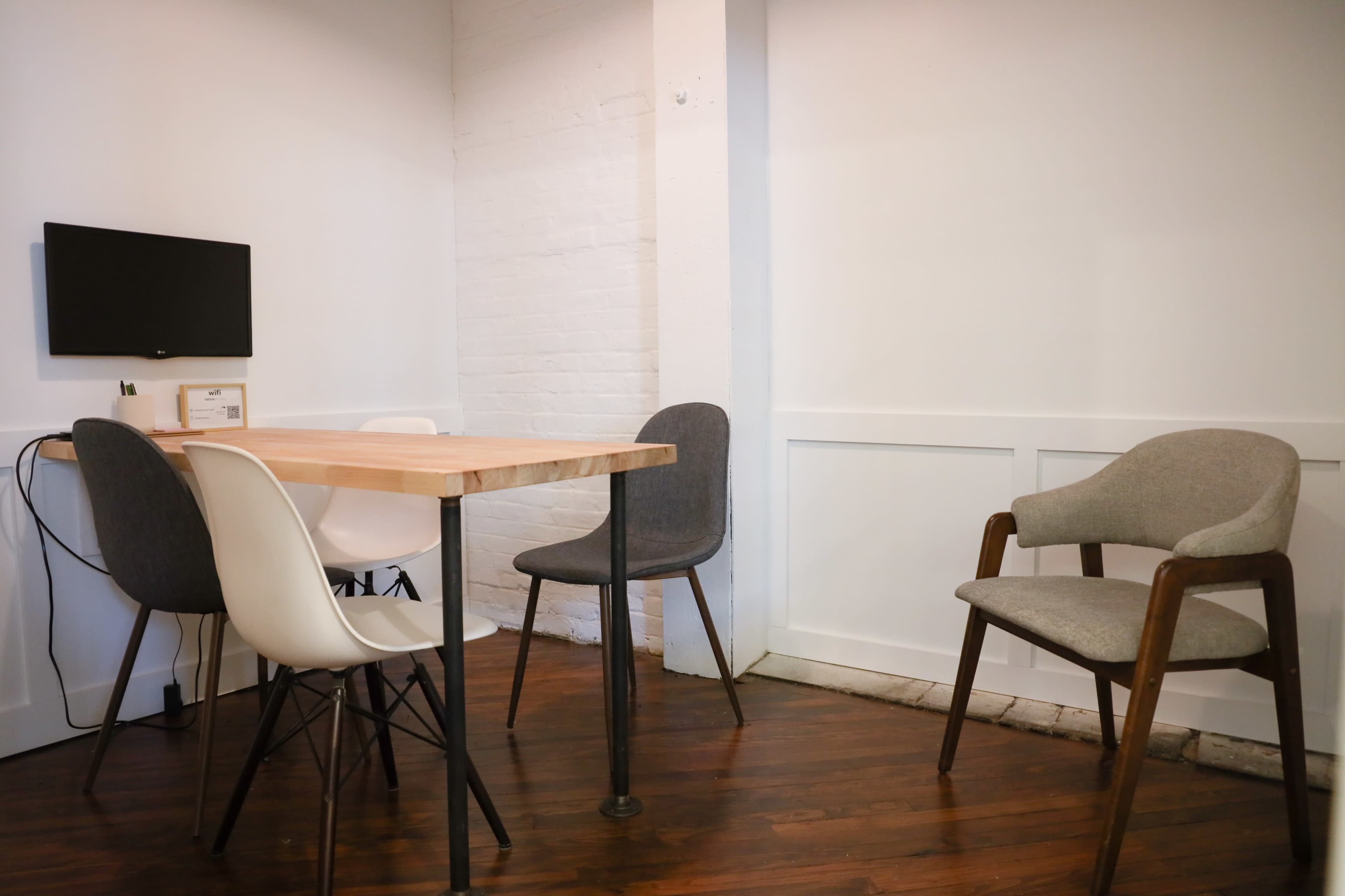 A small, minimally furnished meeting room with a wooden table surrounded by four chairs and a wall-mounted television.