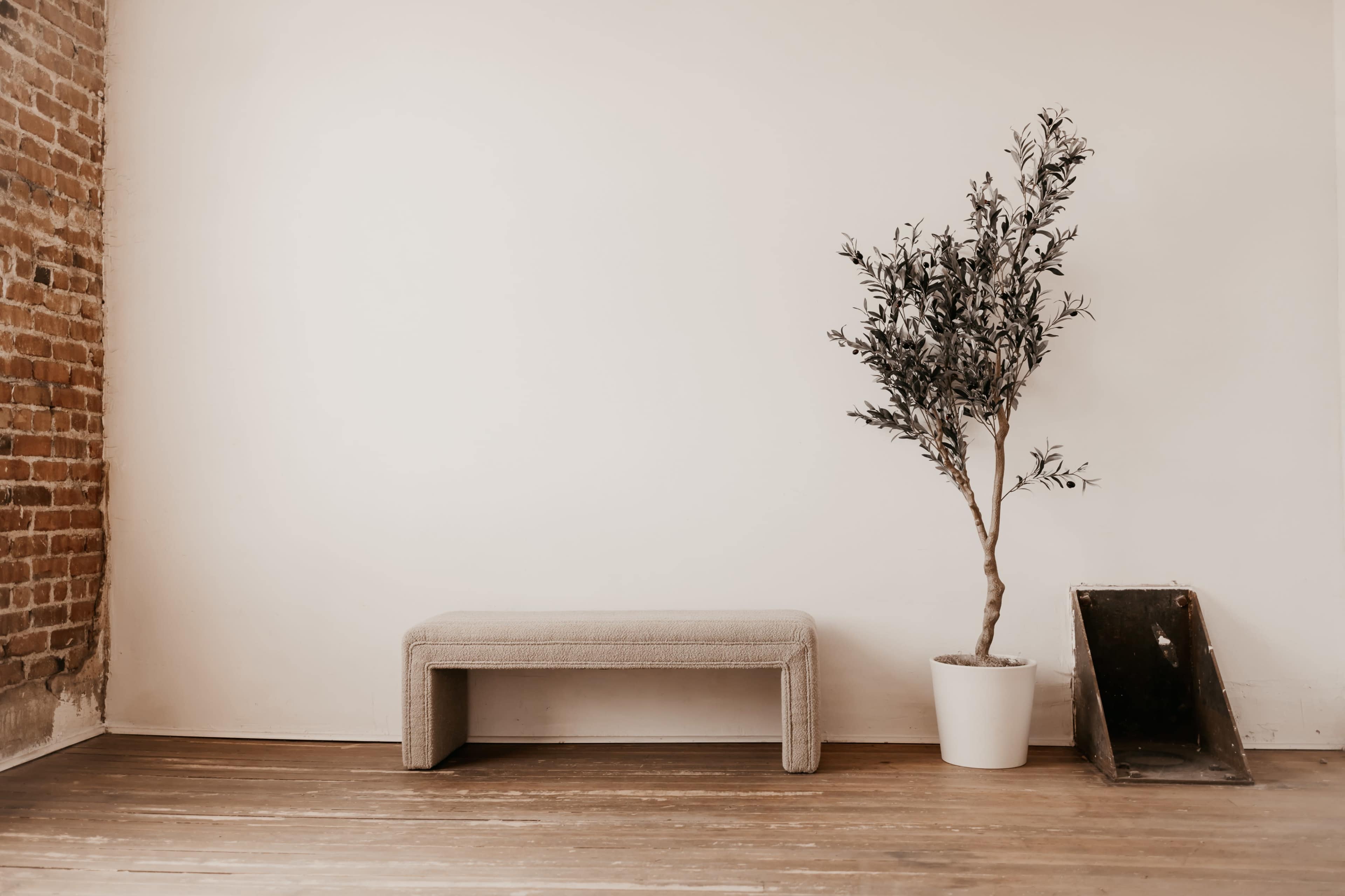 A bench next to a potted tree on a wooden floor, with a brick wall and a plain white wall in the background.
