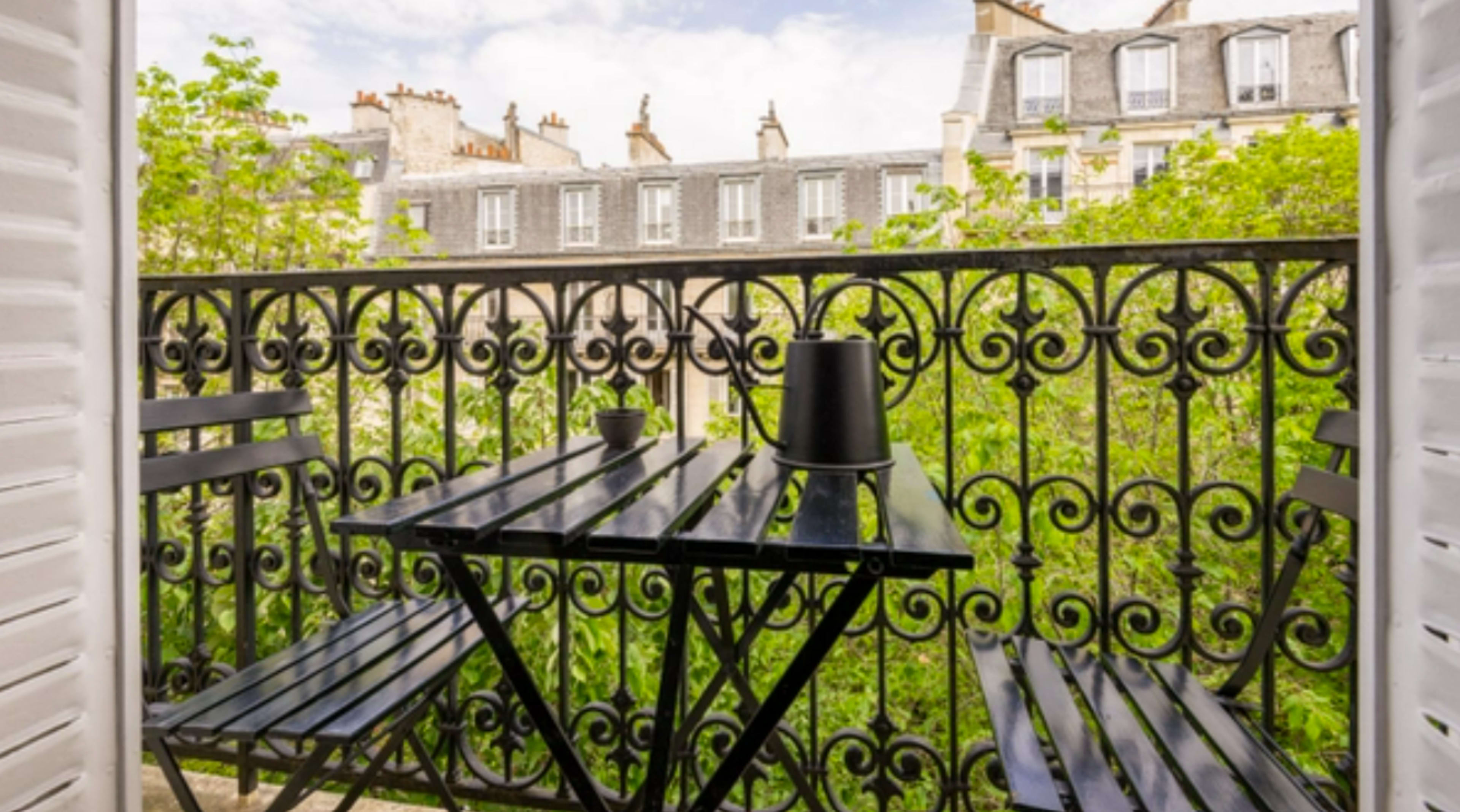 A small black dining table and two chairs are set on a balcony overlooking a tree-filled courtyard and classic Parisian buildings.