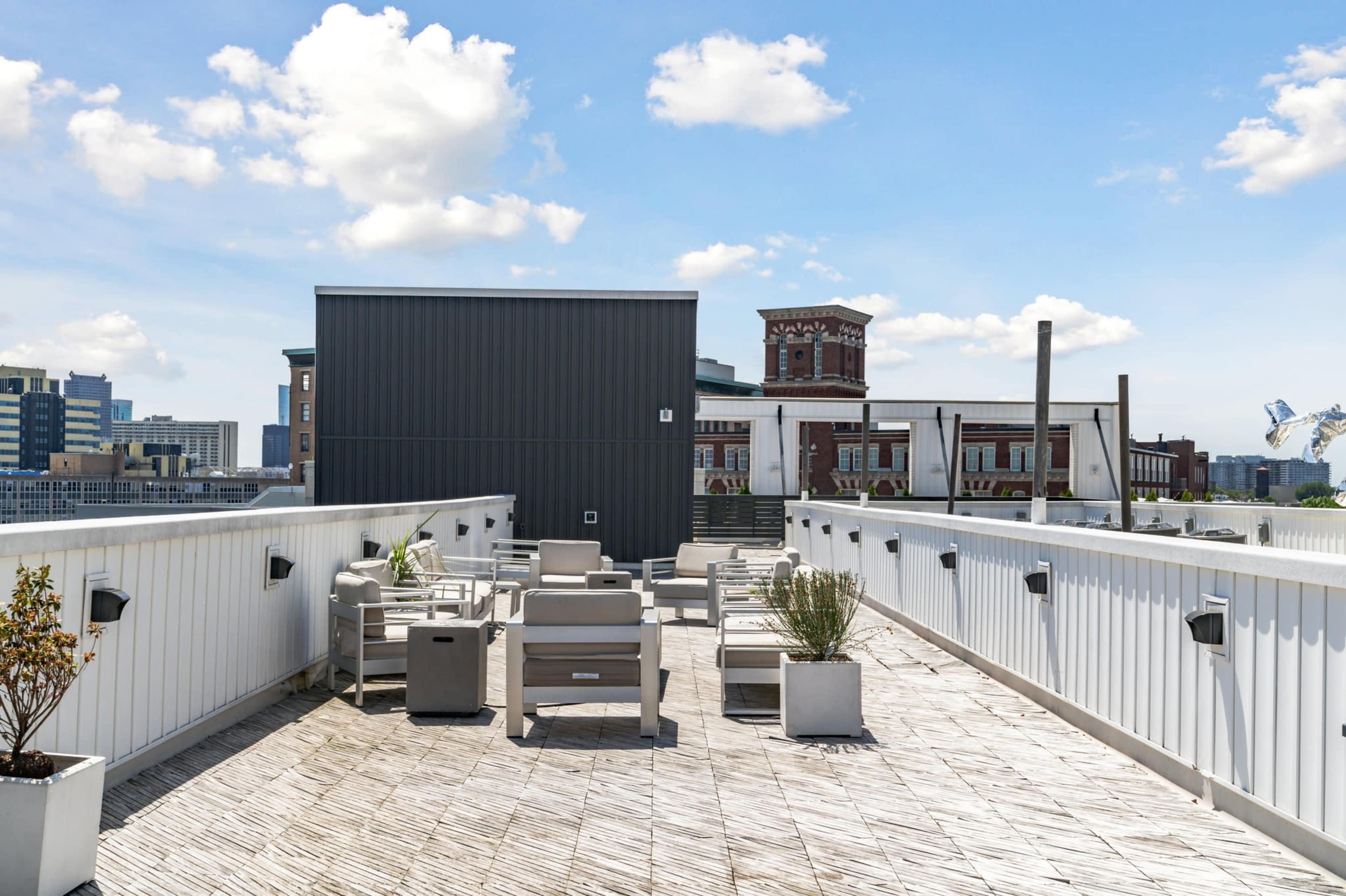 A rooftop terrace with seating areas and planters, overlooking a cityscape against a clear blue sky.
