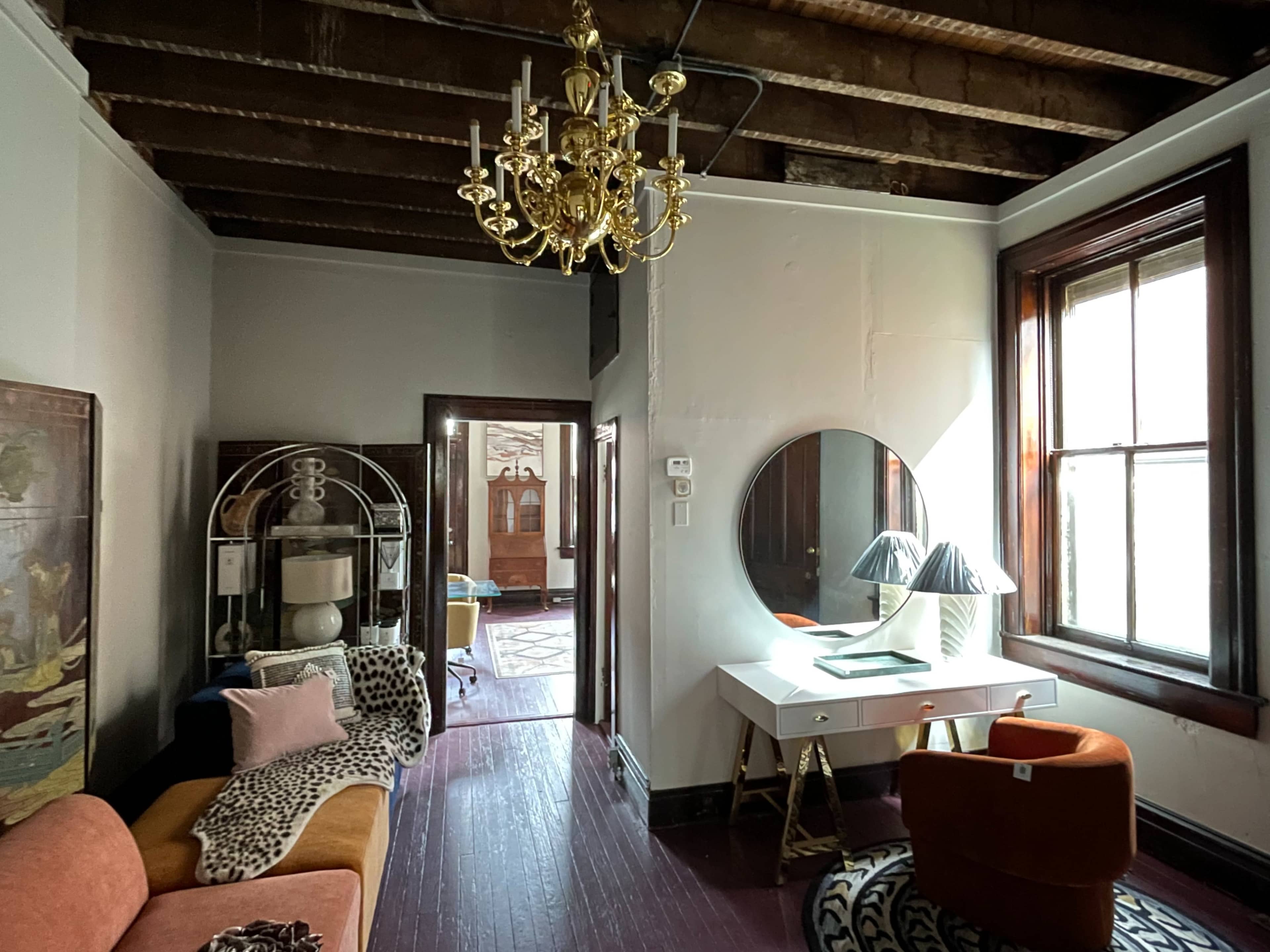 A room with exposed wooden beams, a chandelier, a round mirror above a white desk, and various pieces of furniture including a sofa and an orange chair.