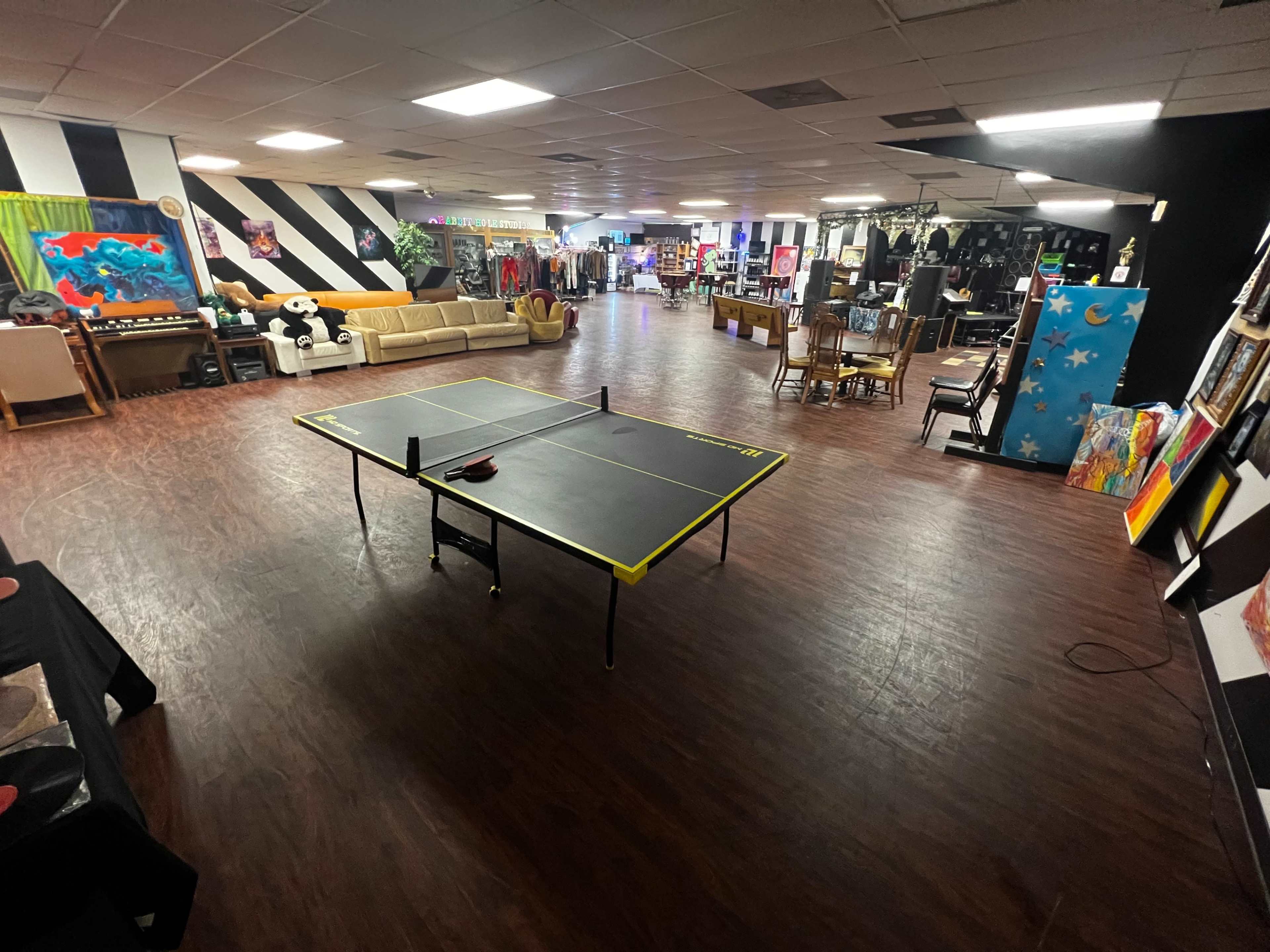 A spacious indoor area featuring a ping pong table at the center, surrounded by various seating options and colorful artwork on the walls.