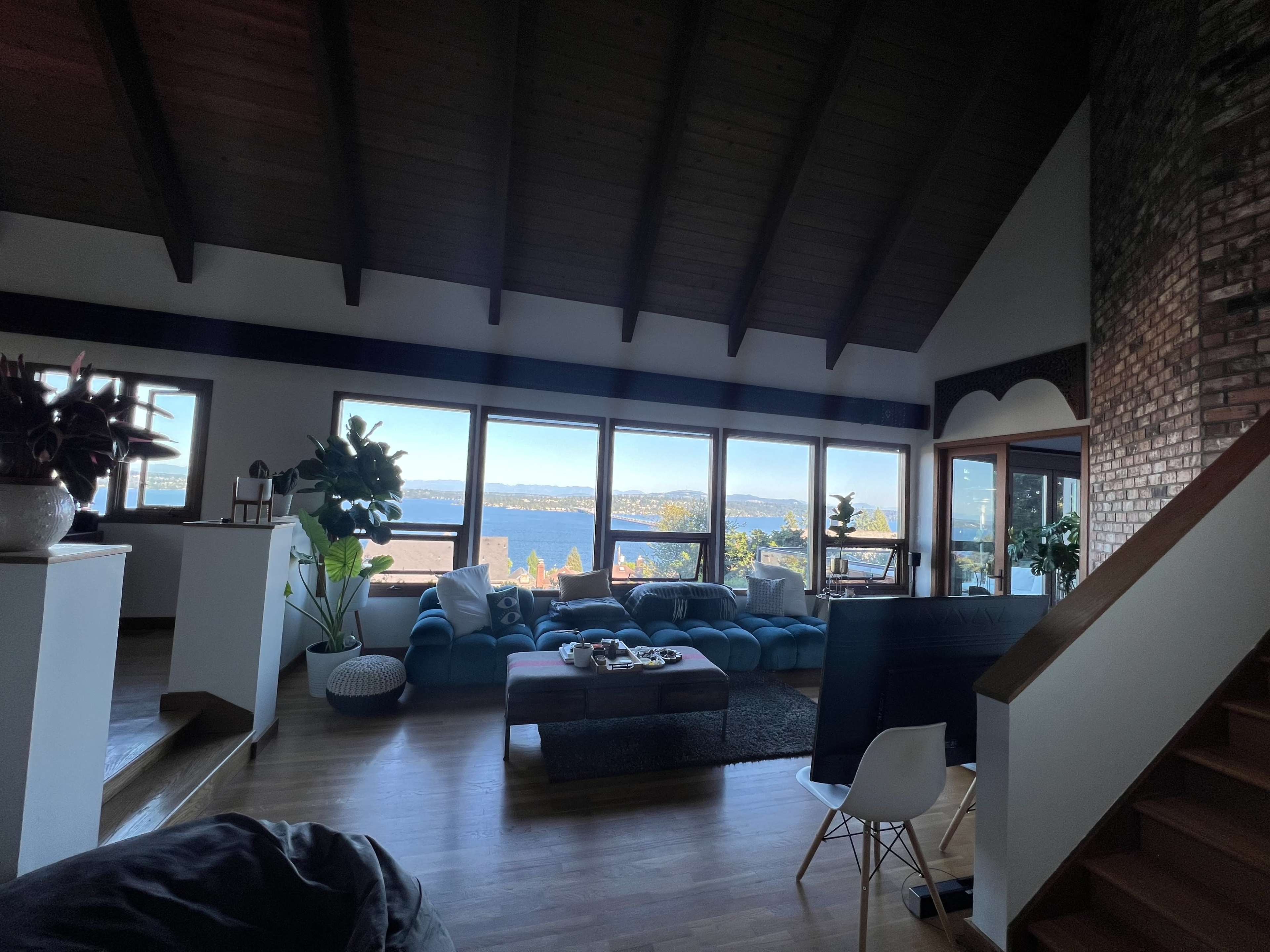 A spacious living room features a blue sectional couch, large windows with a view of the water, and wooden beams overhead.
