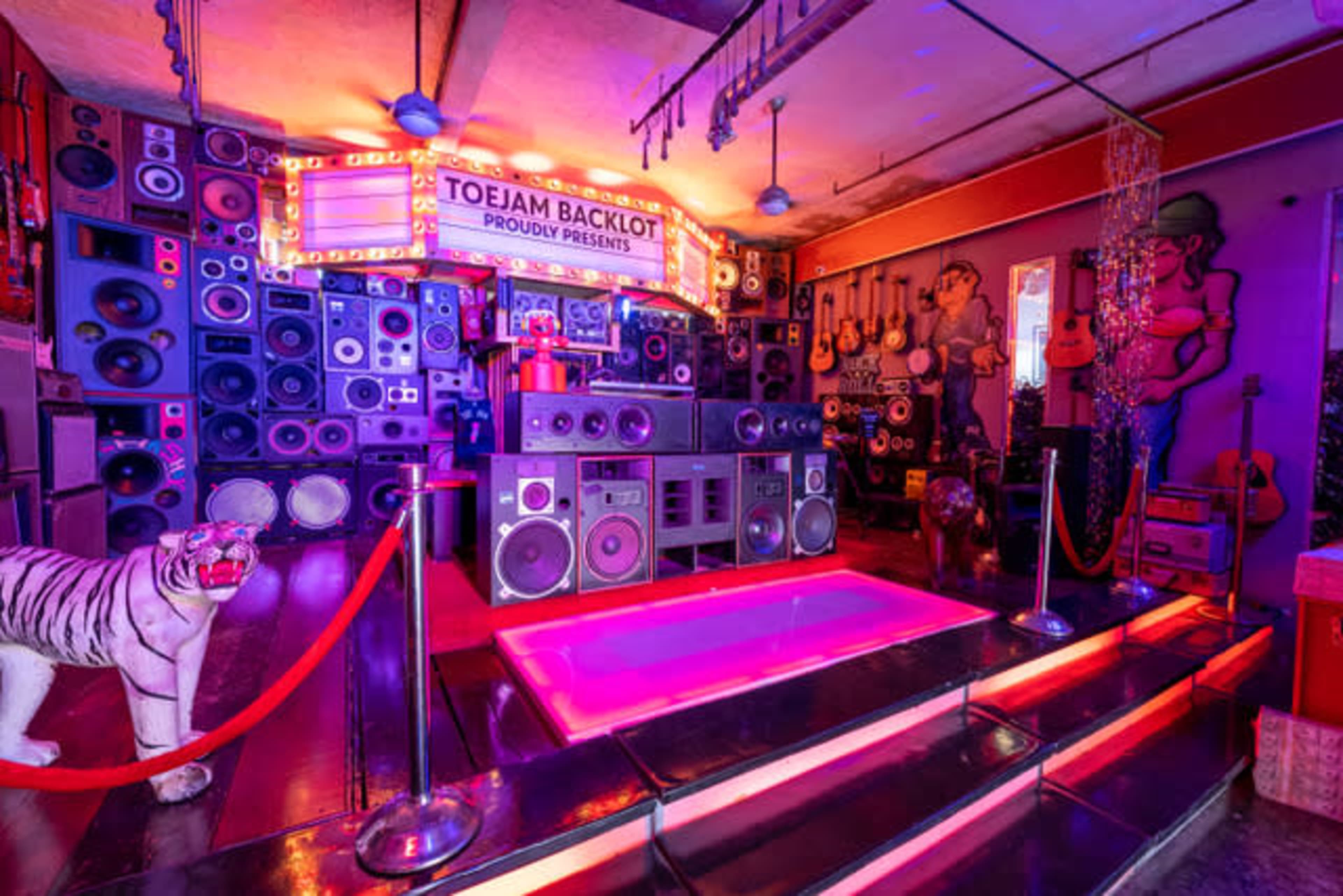 A colorful room filled with an array of speakers, musical instruments, and a pink-lit stage area, complemented by a decorative statue of a tiger and vibrant wall art.