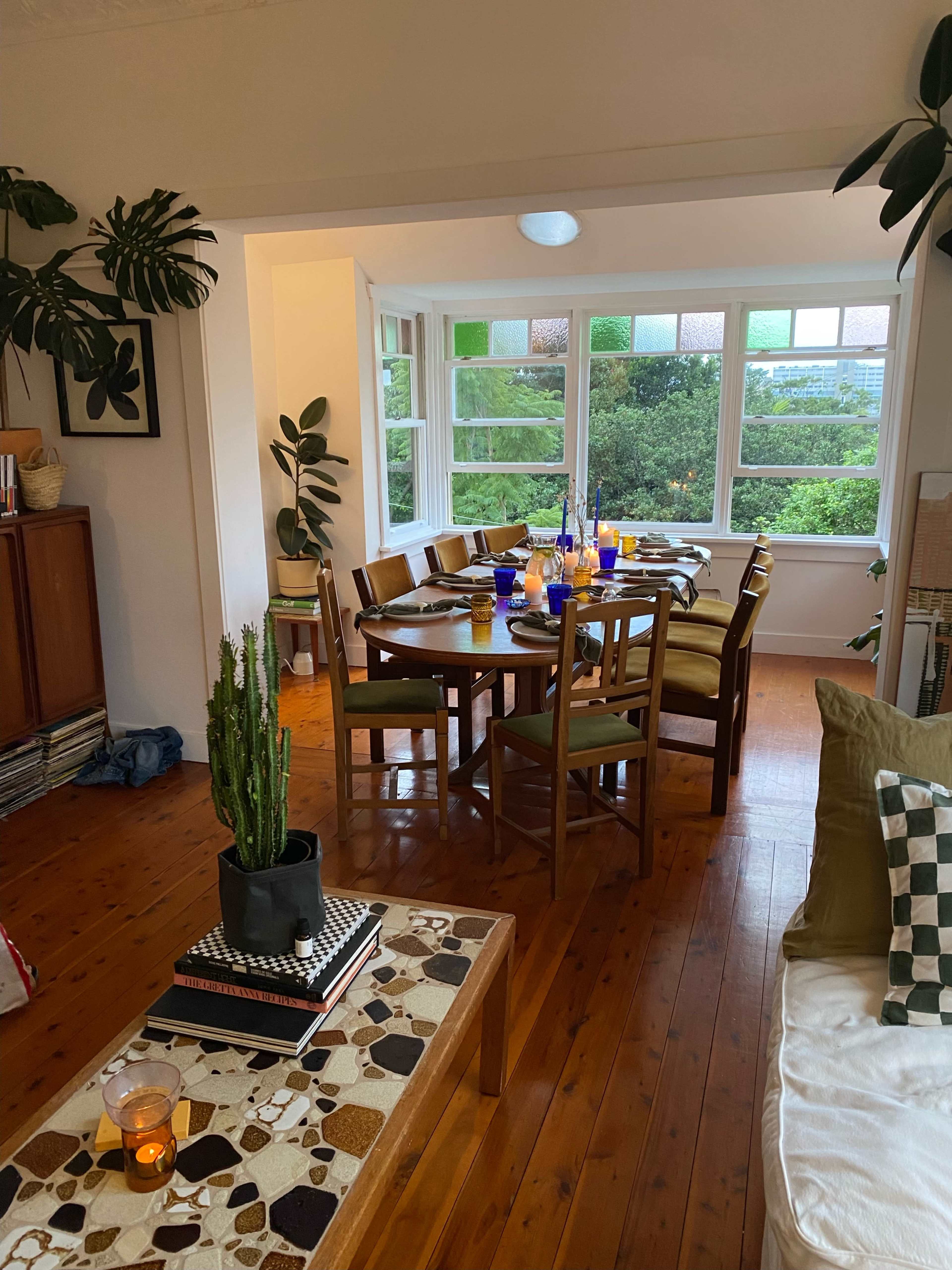 A wooden dining table set for a meal is situated in a well-lit room with large windows and greenery visible outside.
