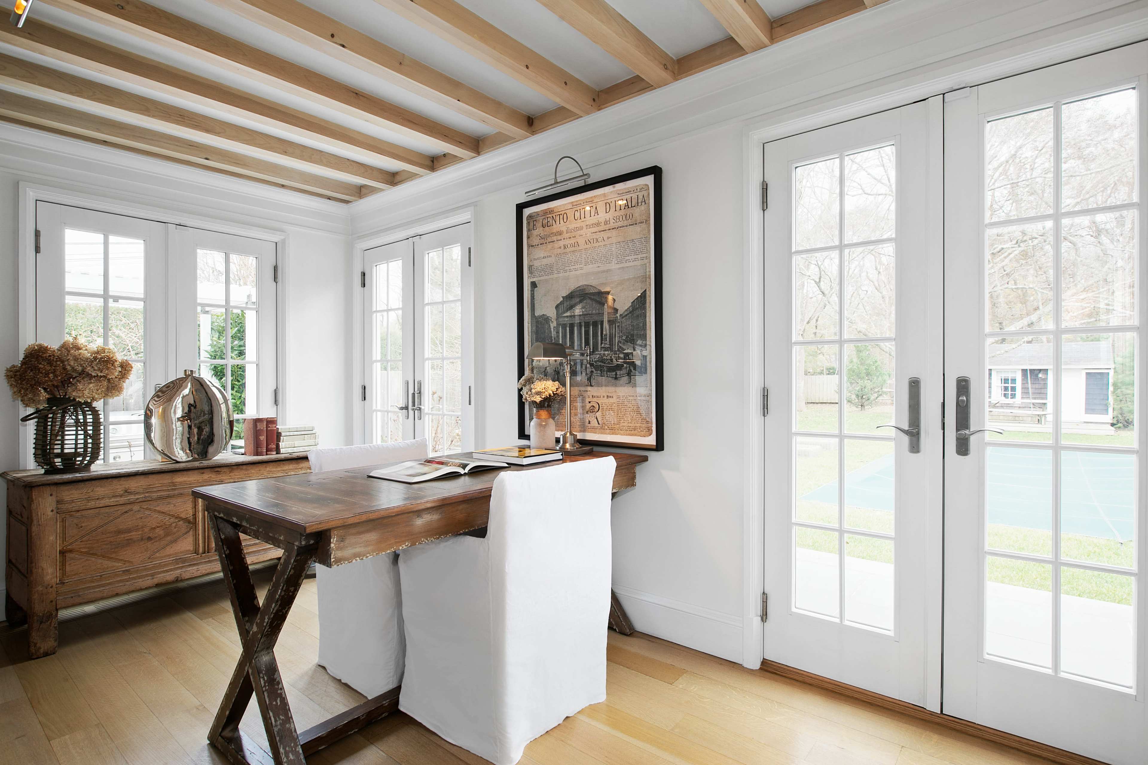 A wooden desk is positioned near large glass doors that open to a garden, surrounded by white walls and exposed wooden beams.