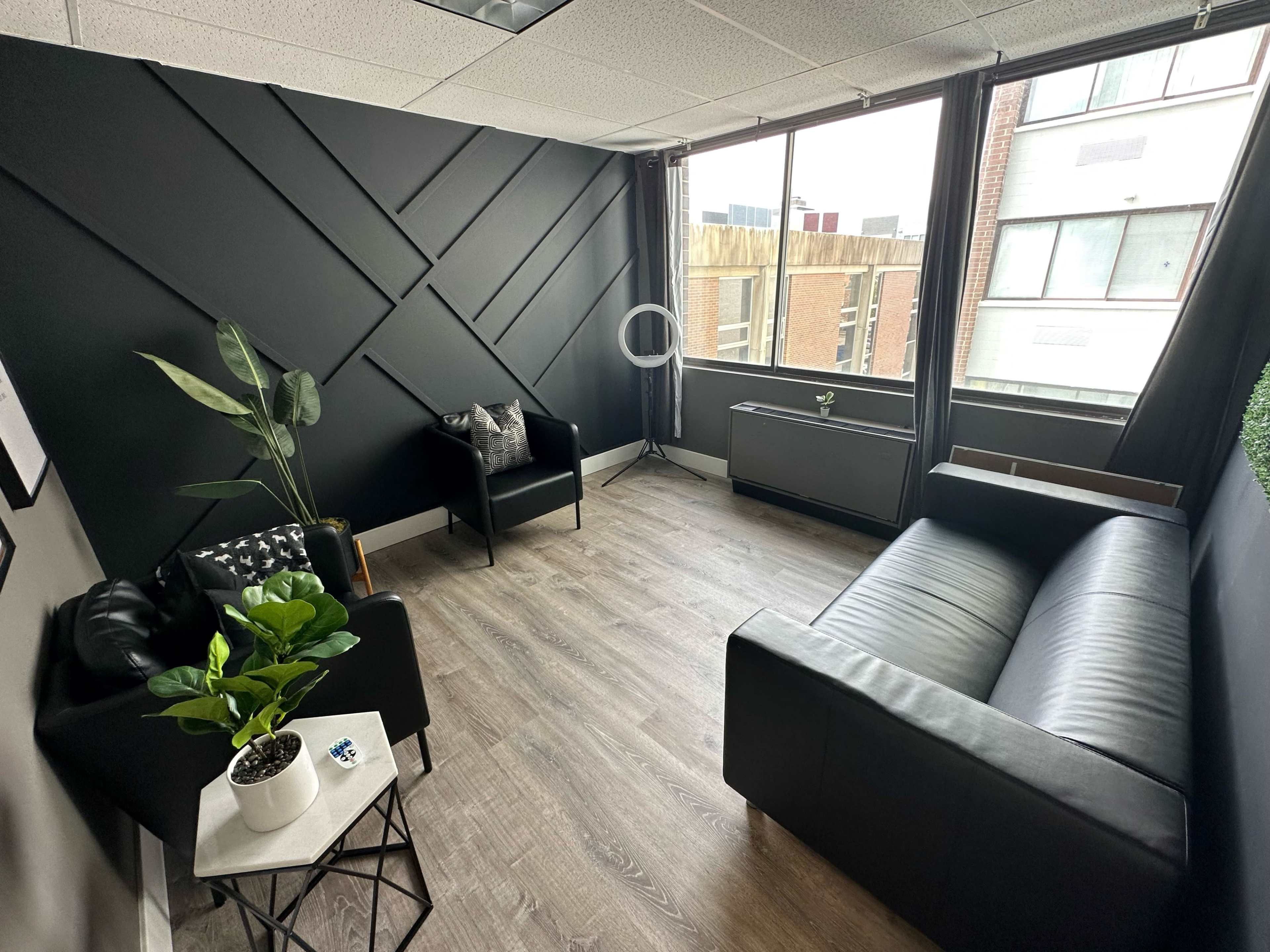 A modern, minimalistic lounge with a black accent wall, a black sofa, an armchair, a small potted plant on a side table, and large windows revealing a city view.