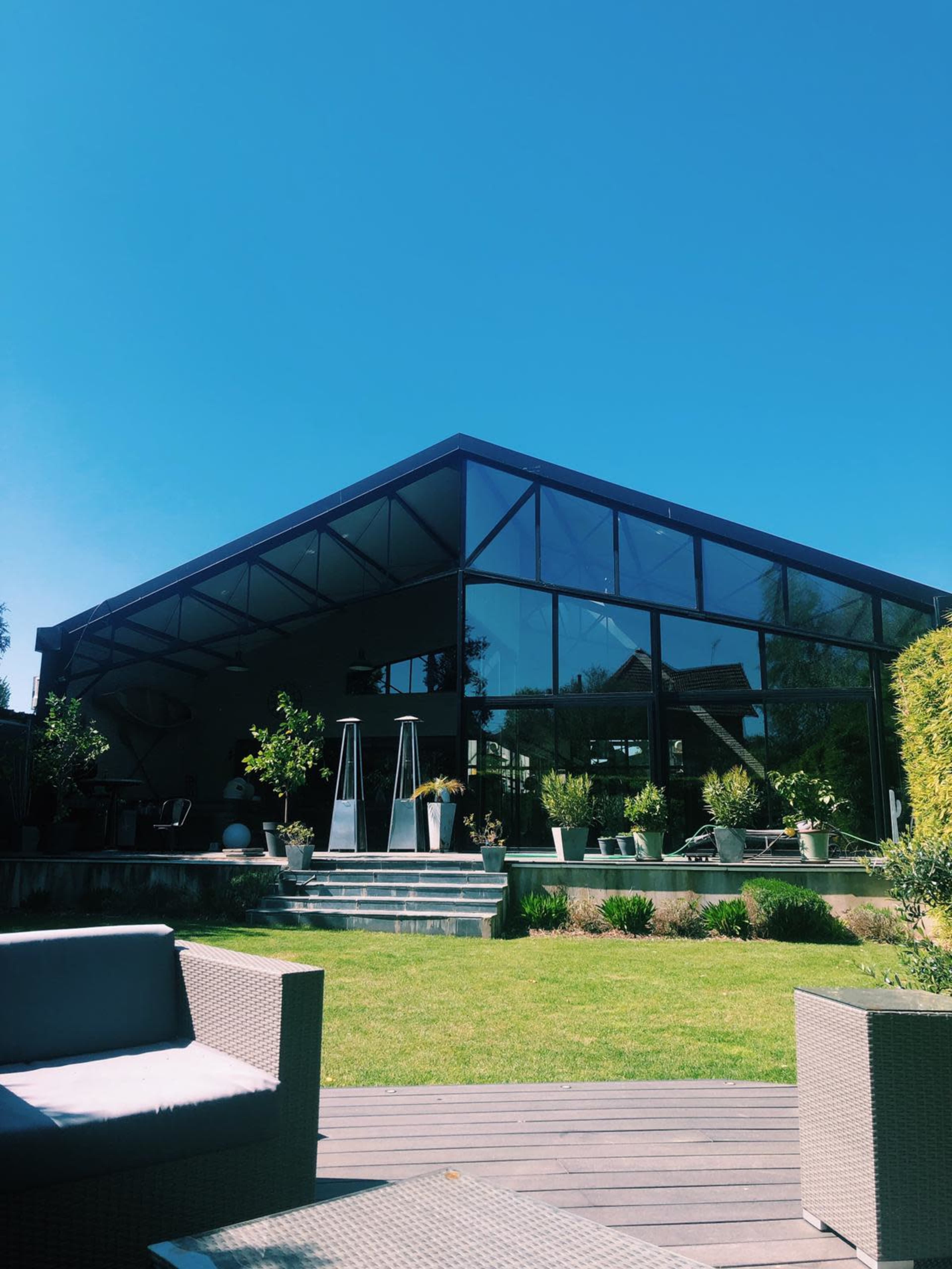 A modern glass-panel house with a sloped roof is surrounded by well-maintained greenery and outdoor seating.