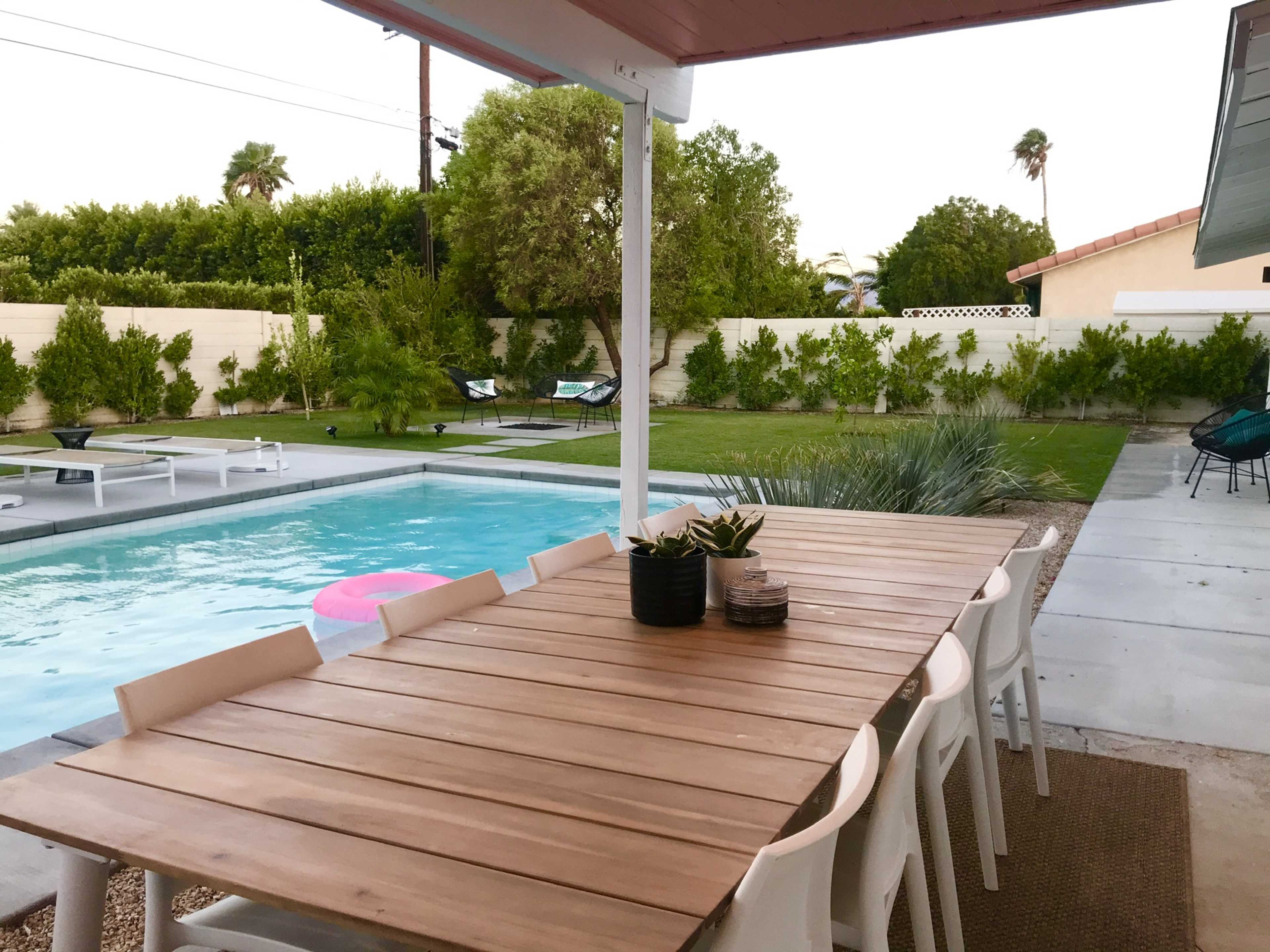 A wooden dining table with white chairs is set beside a swimming pool in a spacious backyard featuring lush greenery and lounge chairs.