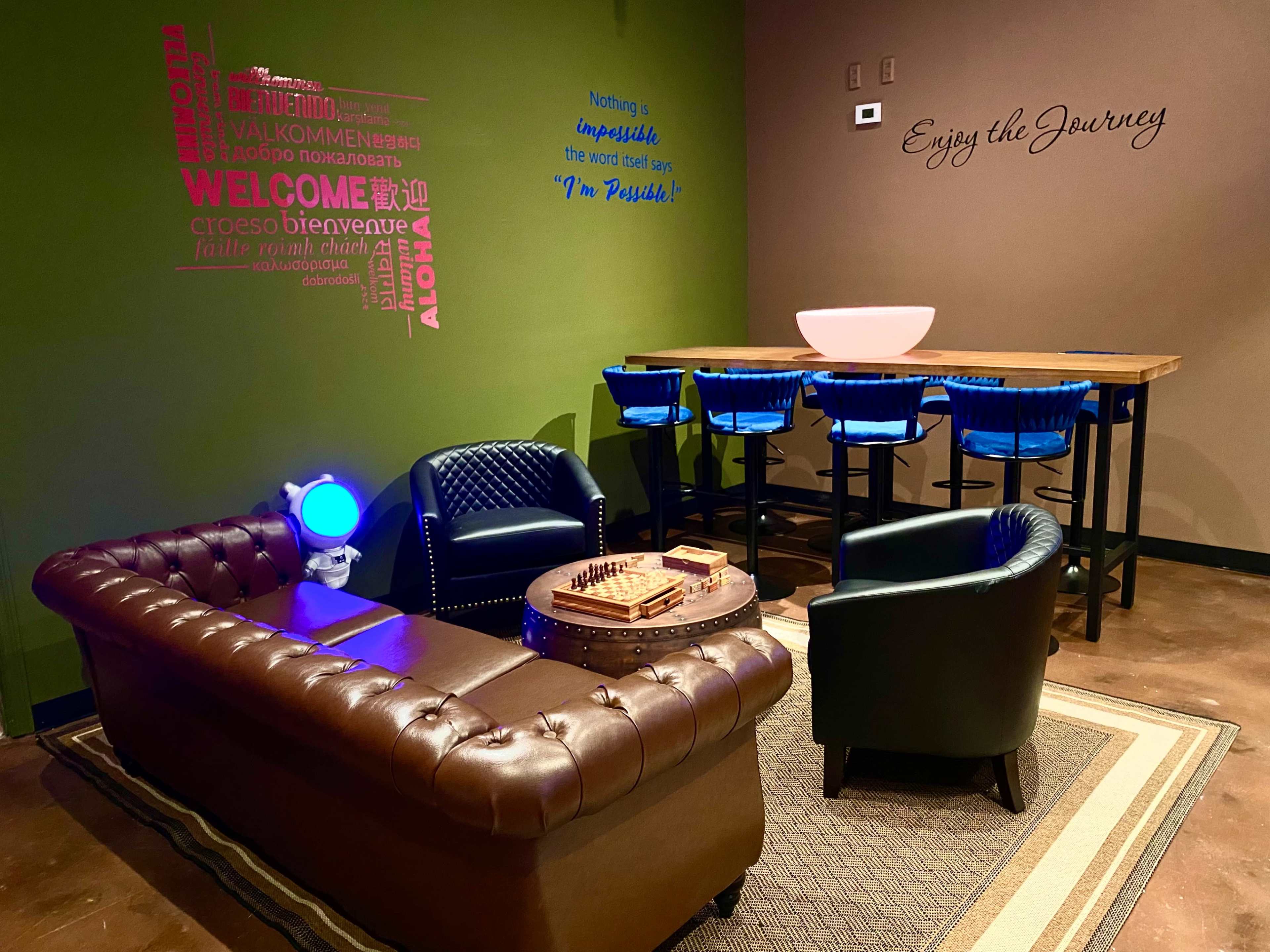 The image shows a cozy lounge area featuring a brown leather couch, a coffee table with a chess set, a bar table with blue high stools, and motivational wall decor.