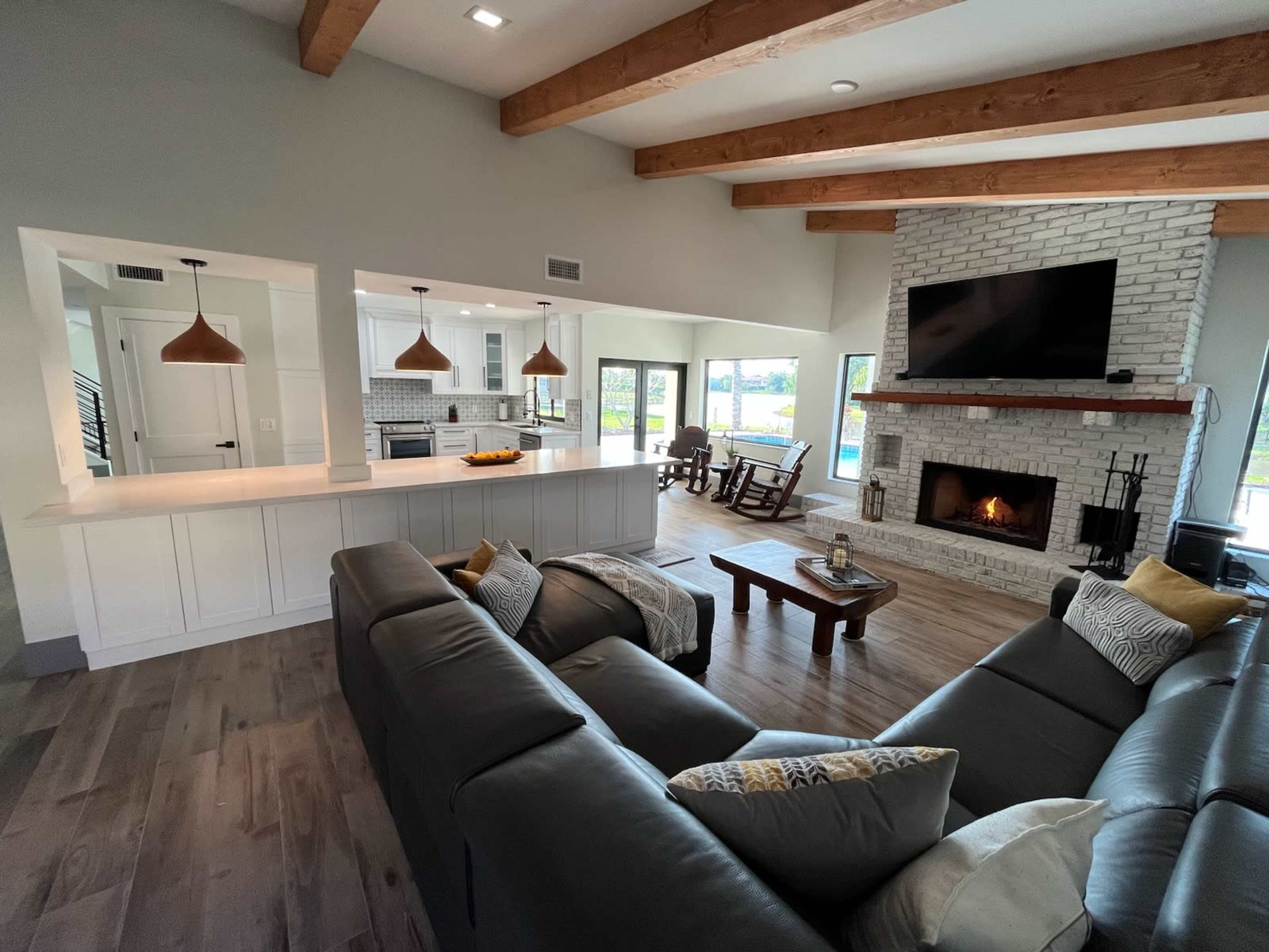A spacious living room features a sectional sofa, a wooden coffee table, and a fireplace, with a view of the kitchen and large windows overlooking a pool area.