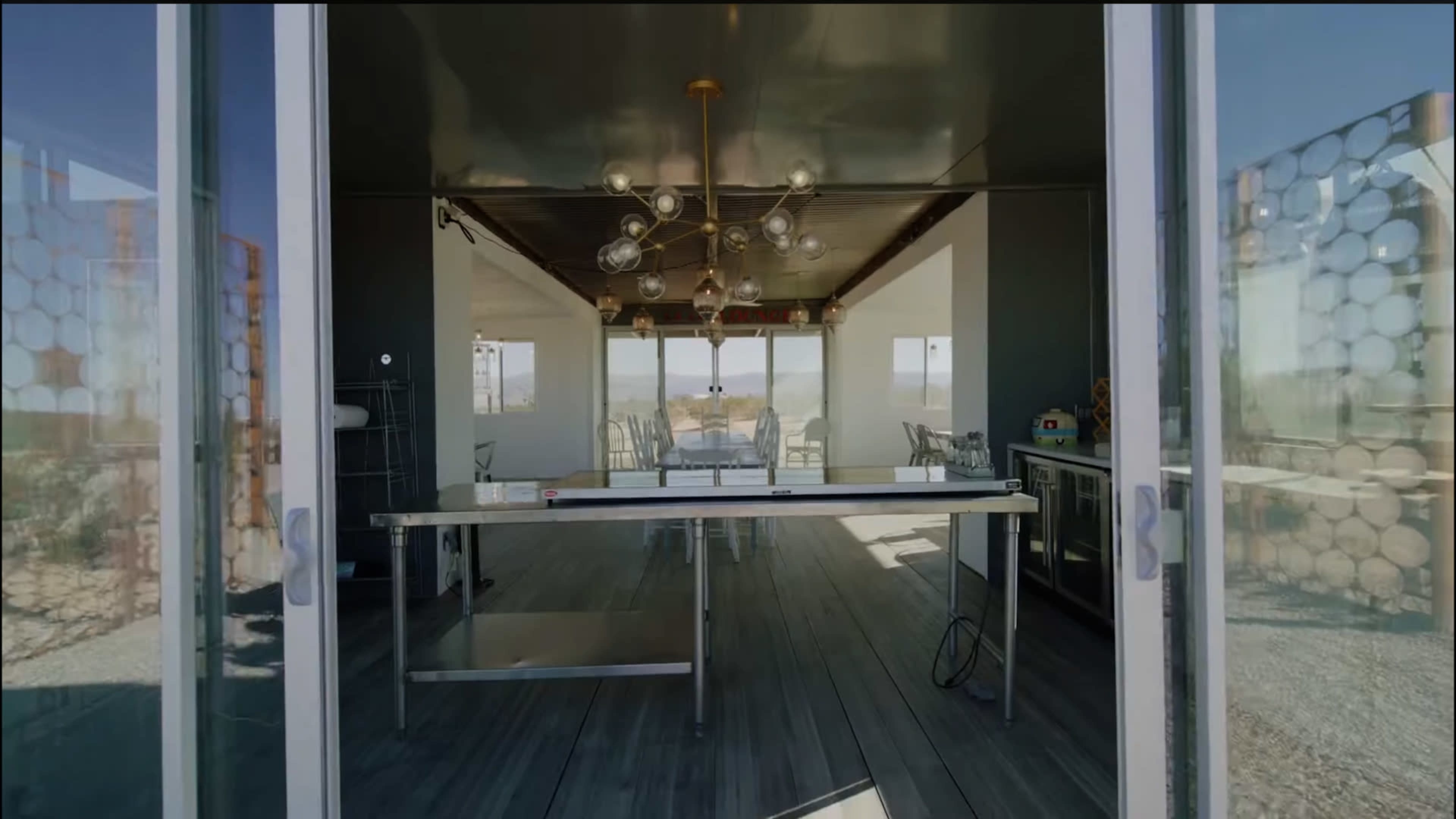 The image shows a modern kitchen area with a glass door opening to a dining space featuring a large chandelier and a view of the outdoors.