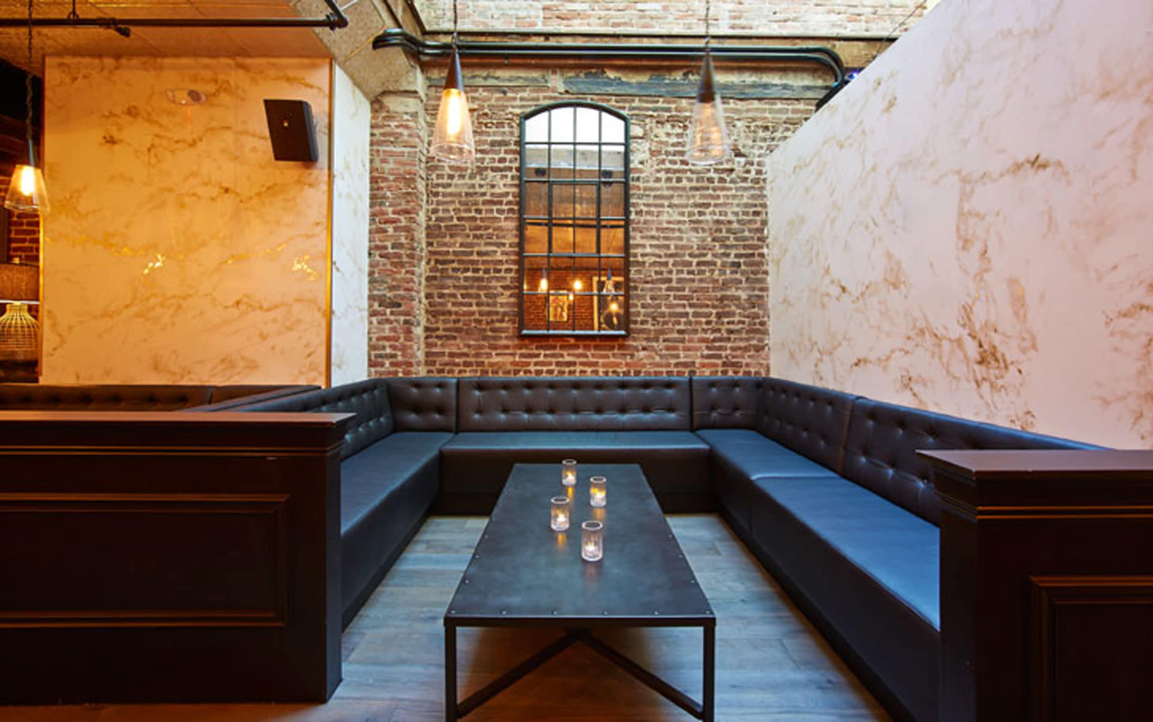A modern lounge area features a rectangular, dark table surrounded by a U-shaped leather banquette and brick walls with large windows.