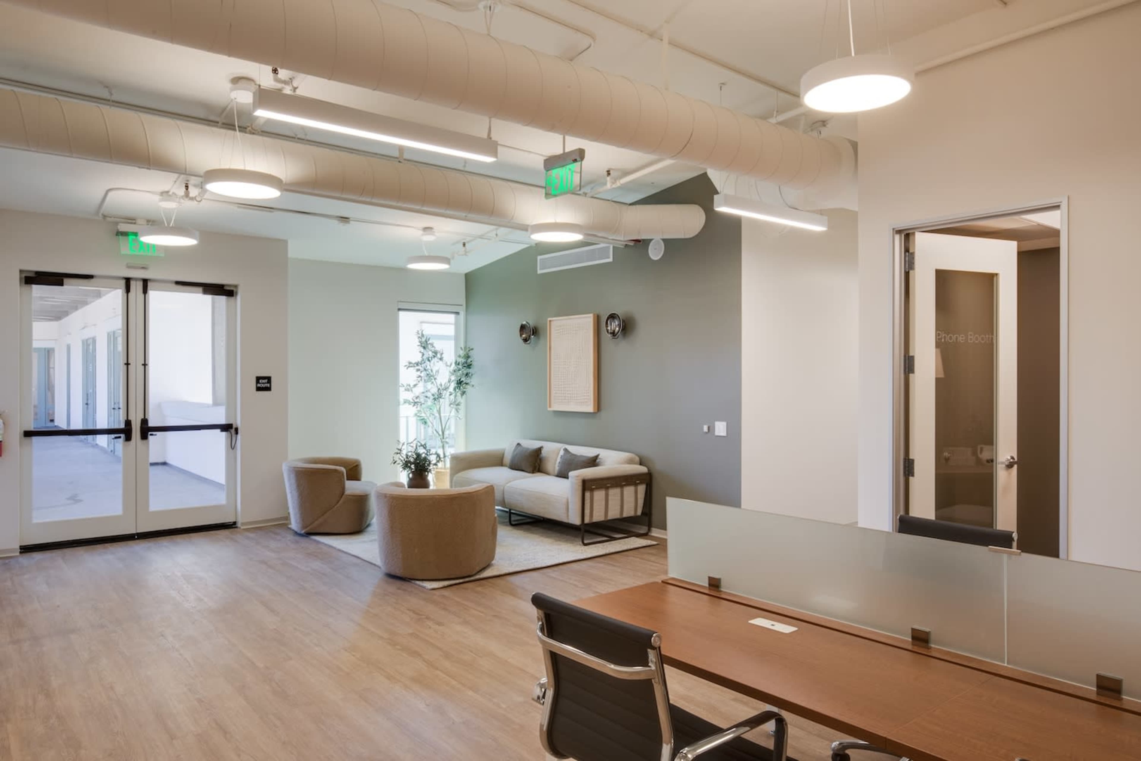 A modern office space featuring a seating area with a sofa and round chairs, a glass partitioned door to a phone room, and natural light entering from large windows.