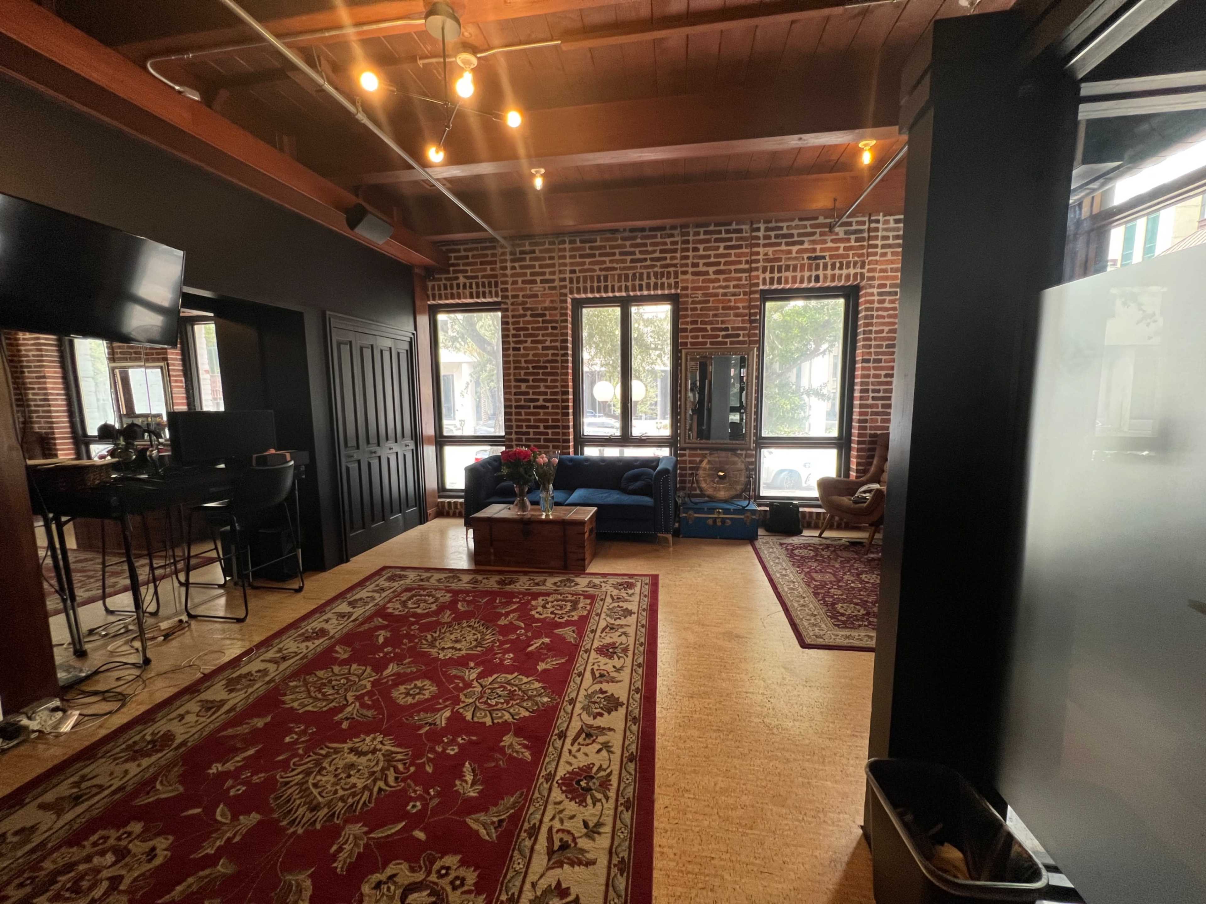 The image shows a modern interior space with exposed brick walls, wooden beams, and two distinct areas featuring furniture, a large rug, and windows.